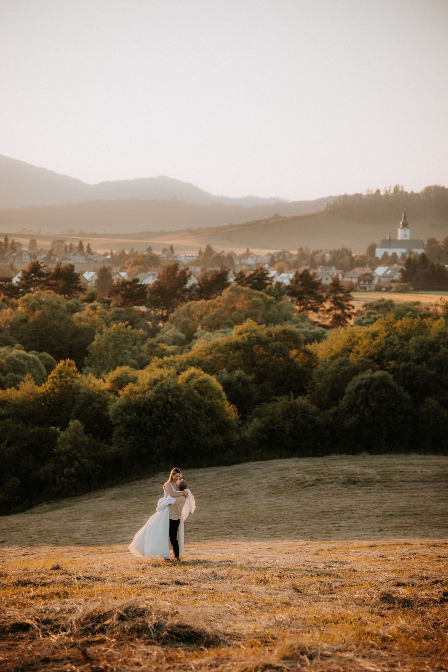 Bride and groom dancing on golden hillside meadow with scenic village, trees, and mountains in soft sunset light.