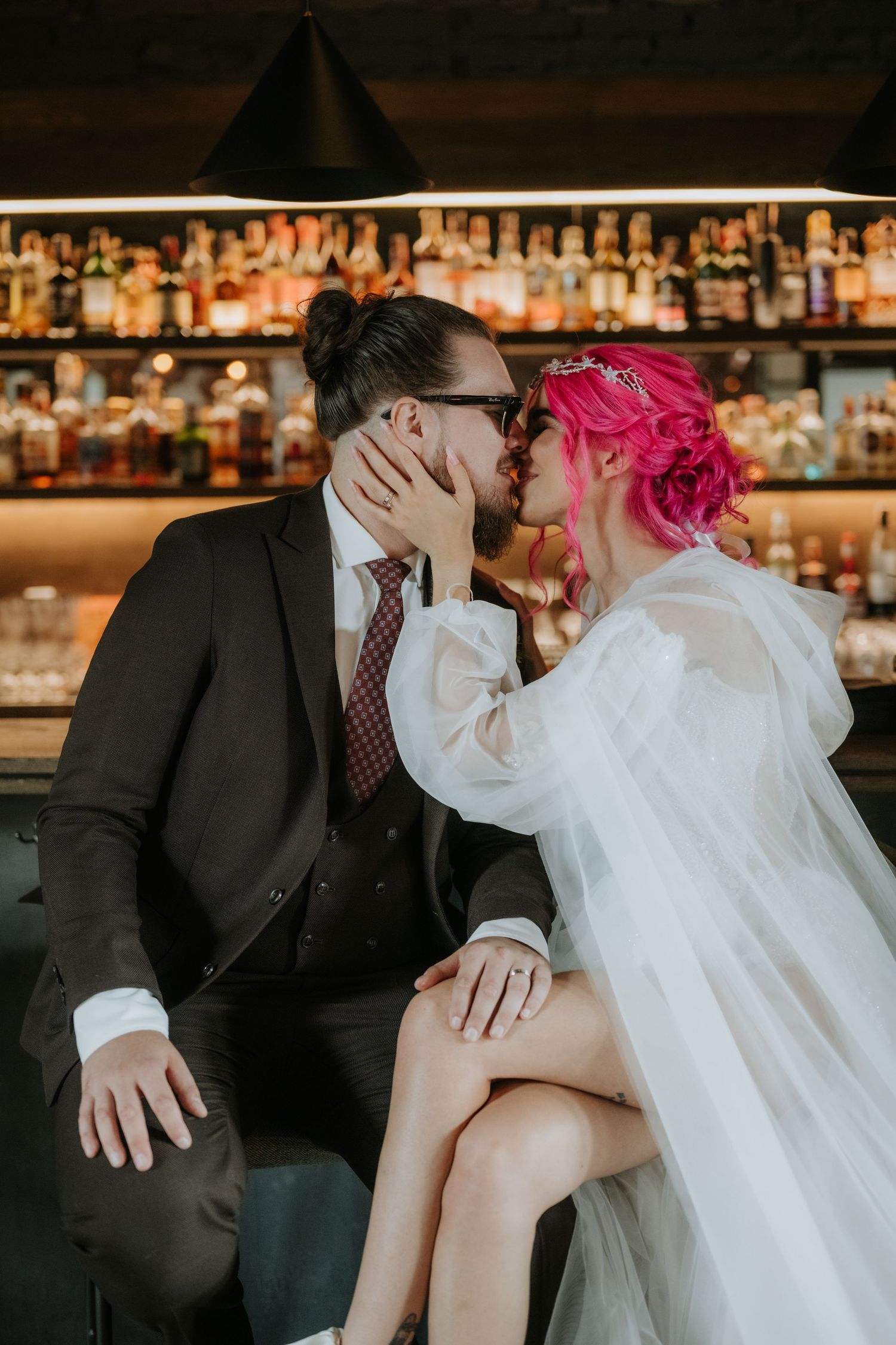 A bride with pink hair and groom in black suit share a kiss at a bar, with colorful bottles on shelves behind them.