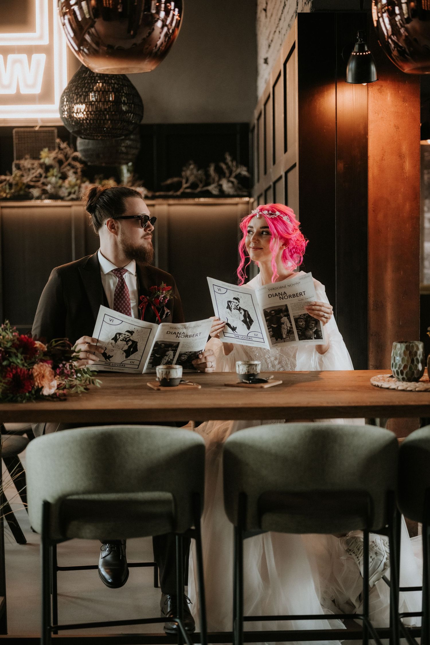 A couple in wedding attire reads newspapers at a wooden table, the bride with pink hair and groom in a dark suit.