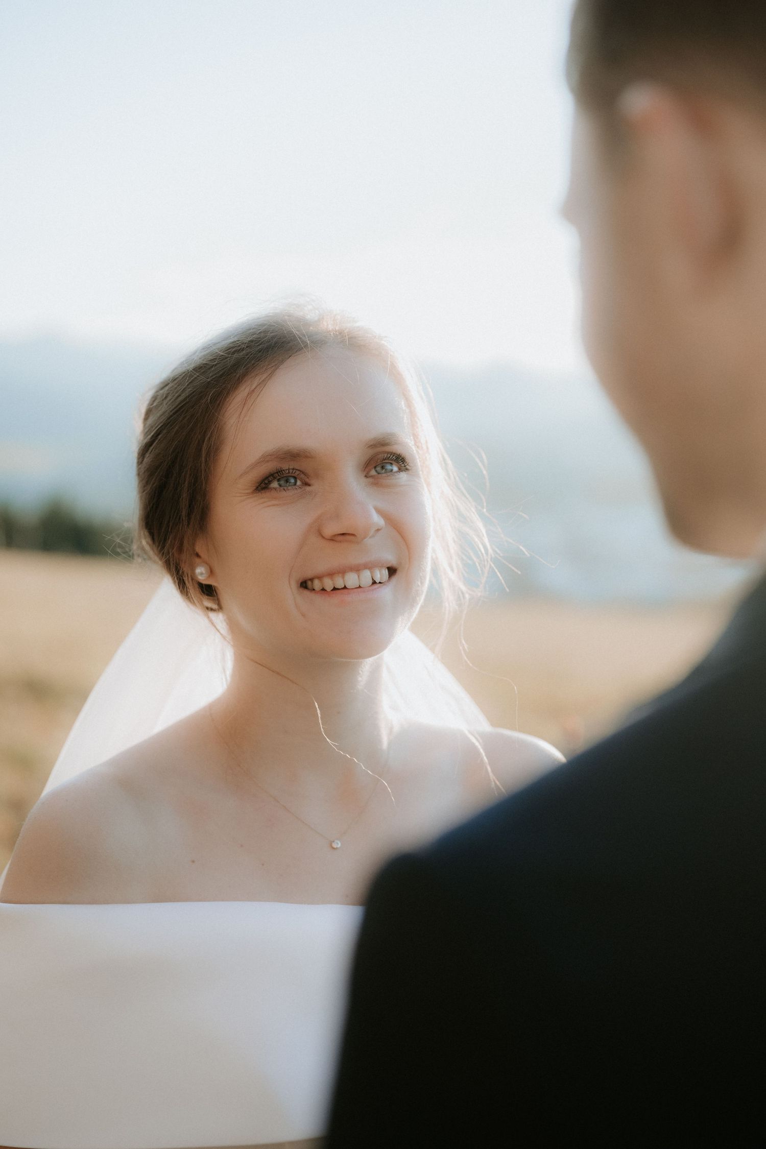 Smiling bride in white dress and veil gazes lovingly at groom during outdoor wedding ceremony with mountain backdrop.