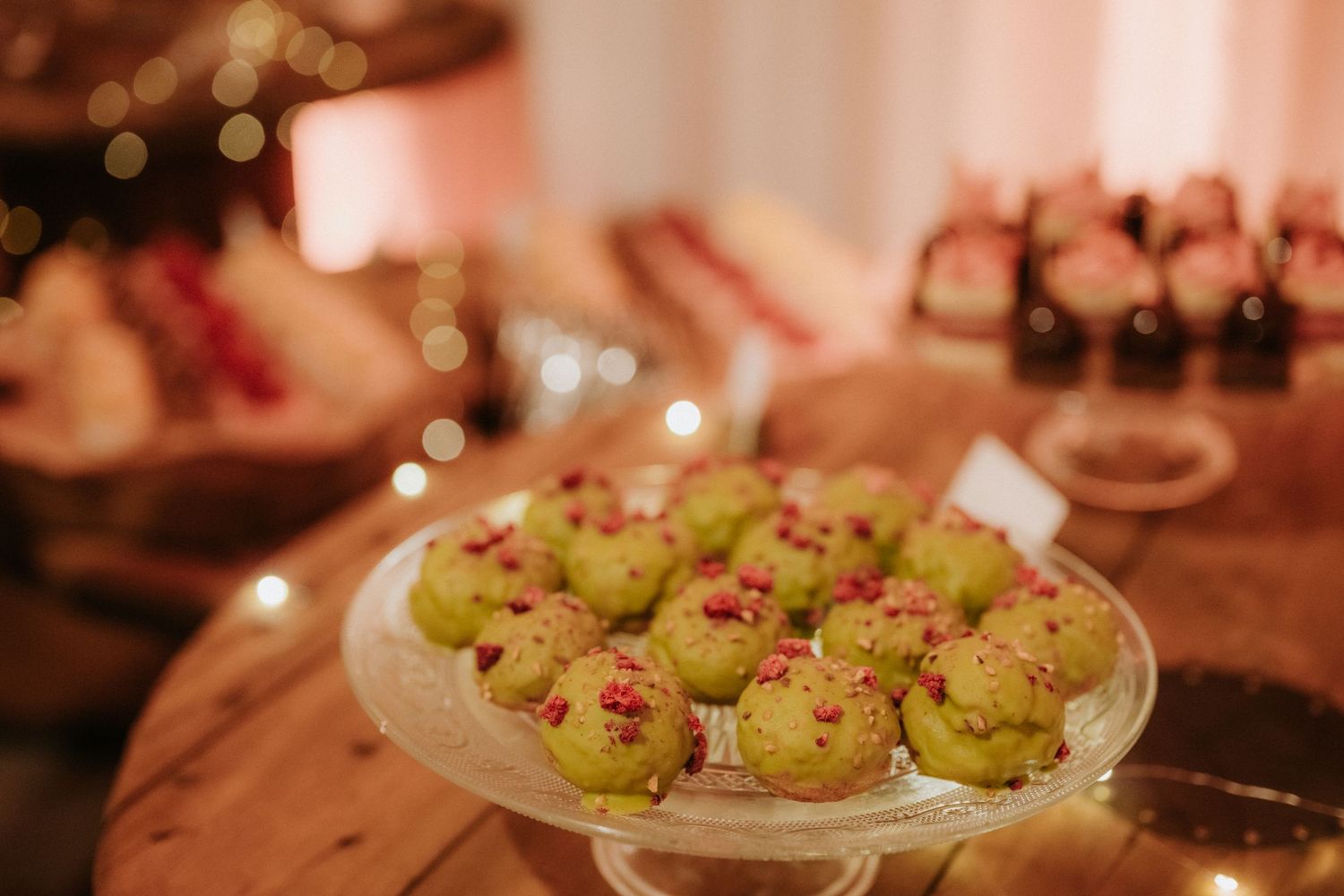Green matcha cake pops topped with red pomegranate seeds displayed on a glass cake stand at an elegant event.