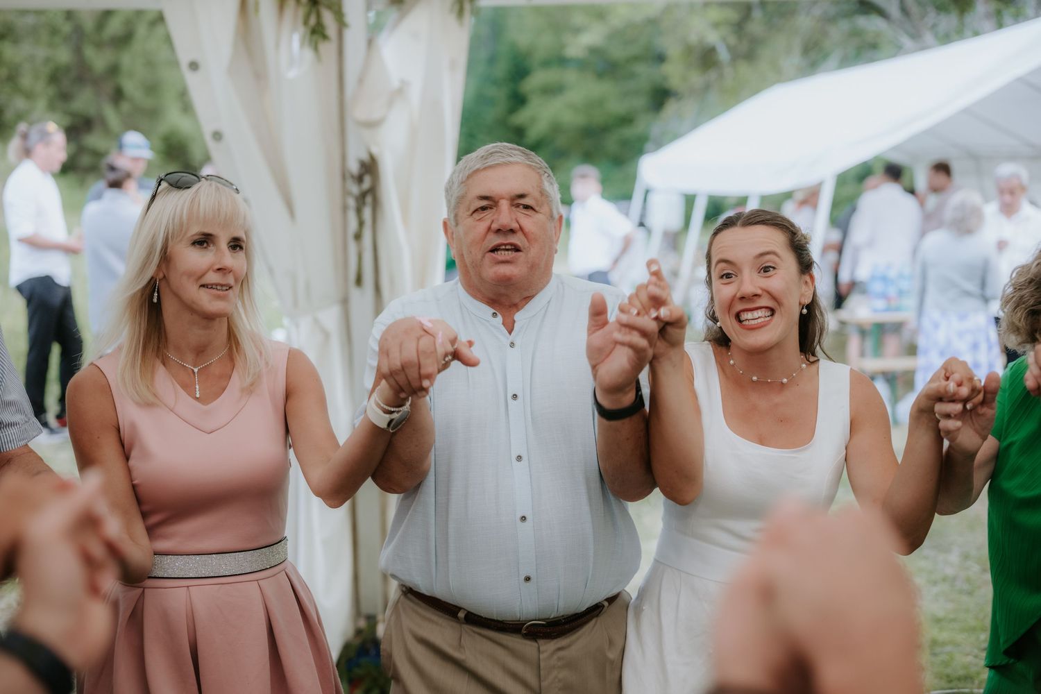 Three guests smiling and dancing at an outdoor wedding reception, wearing formal attire.