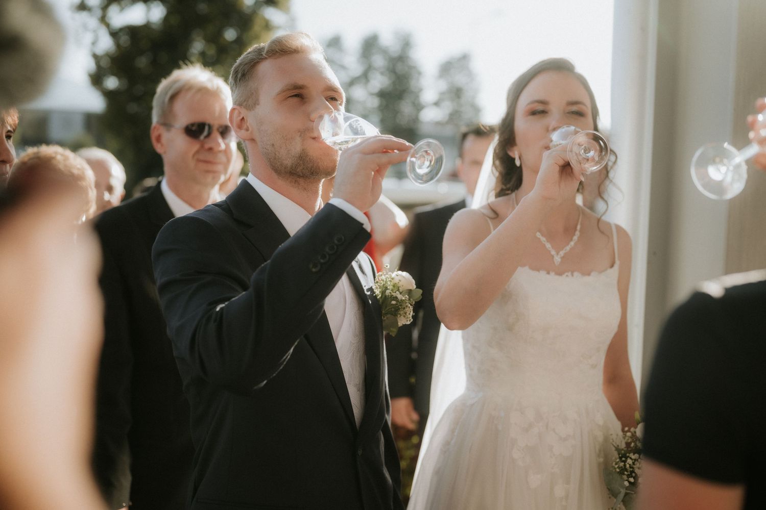 Bride and groom toasting with champagne glasses at outdoor wedding ceremony, guests visible in background.