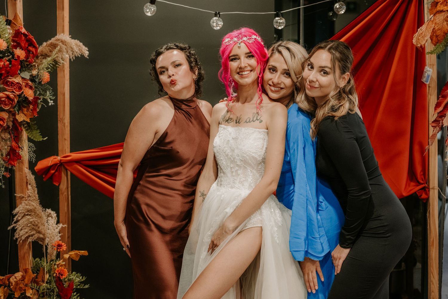 Four women pose together at a wedding, including a bride in white and a guest with pink hair, against a colorful backdrop.