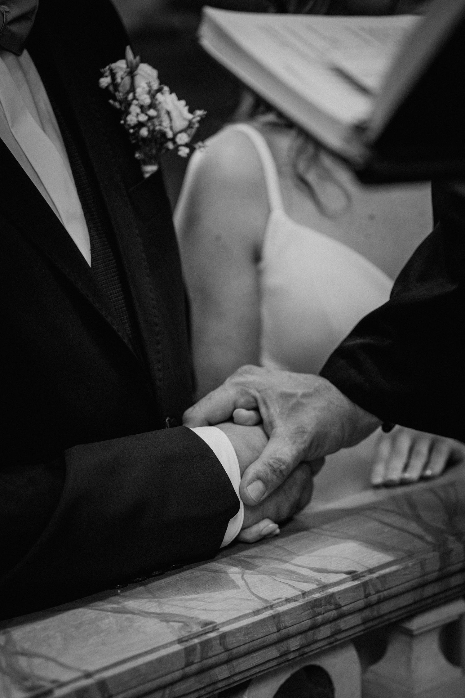 Black and white wedding photo of groom and guest holding hands, bride in white dress visible in background.