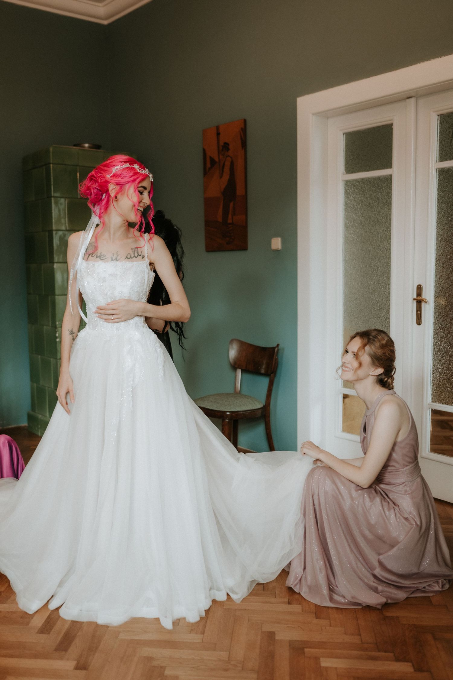 Bride in white gown with pink hair being helped by bridesmaid in mauve dress in elegant green room.