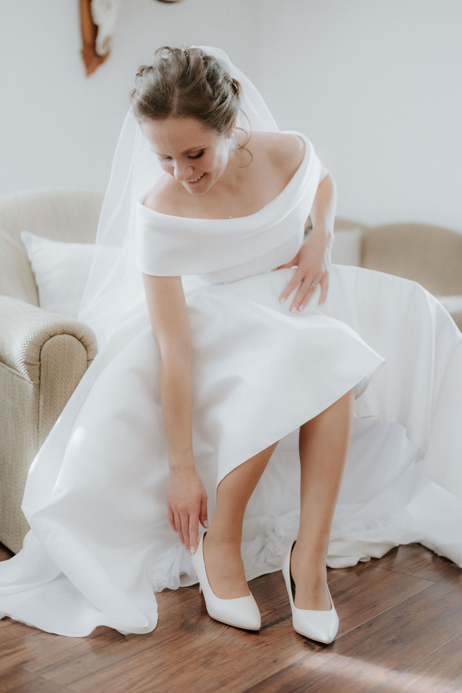 Bride in white off-shoulder gown and veil putting on white heels while seated in a chair.