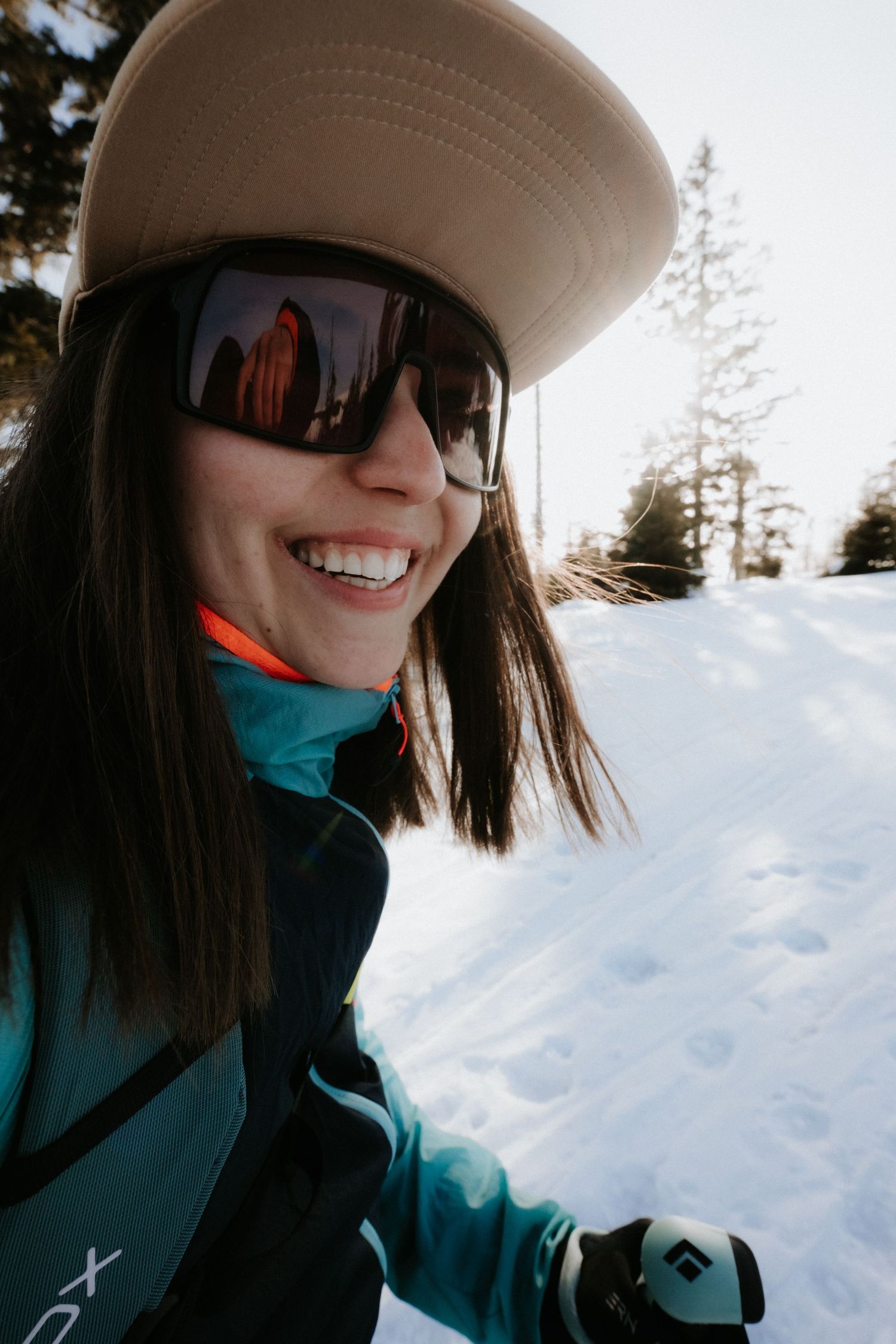 Smiling woman in teal ski jacket, tan cap, and black sunglasses enjoying a sunny winter day on a snowy mountain.