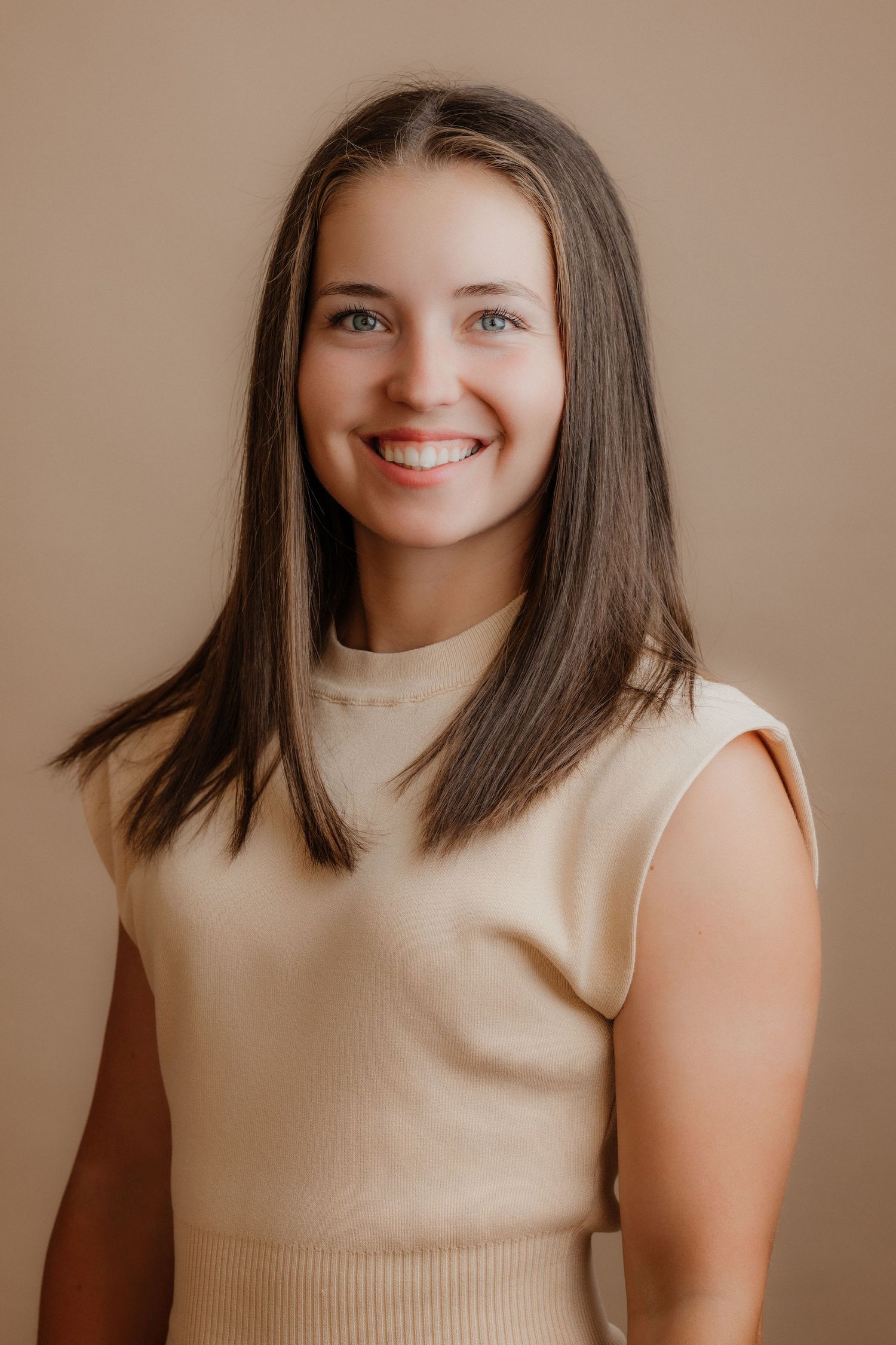 Cheerful young woman with brown hair smiling broadly in cream sleeveless ribbed top against beige background.