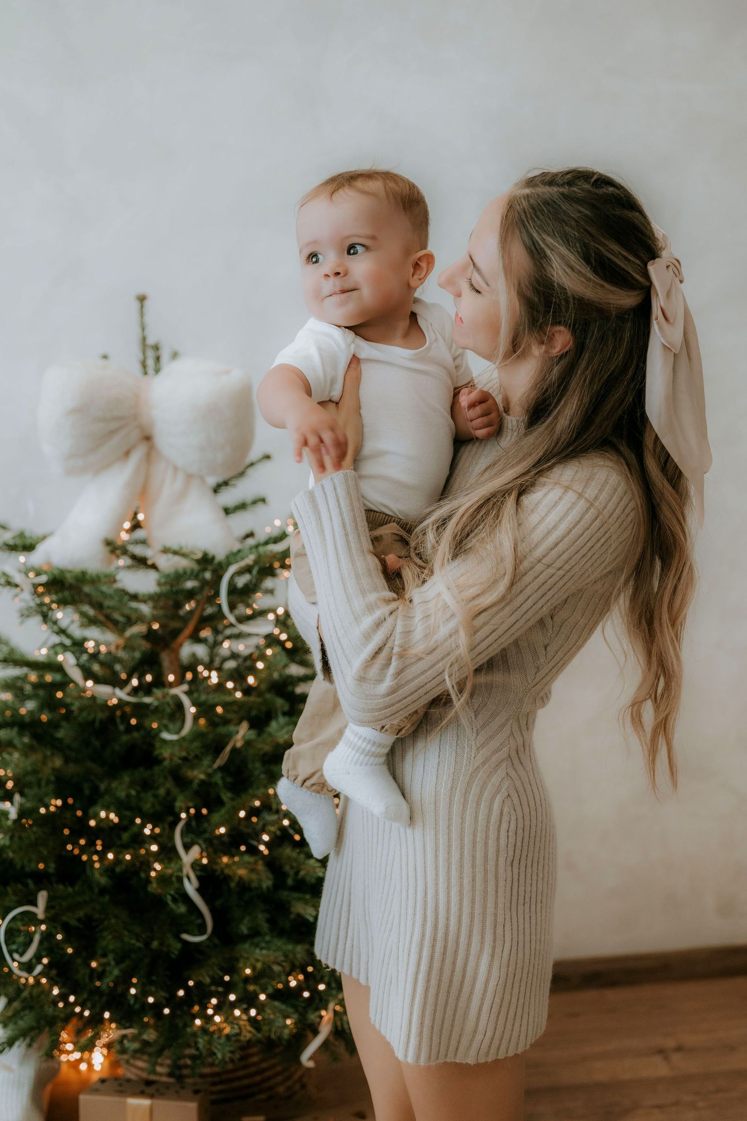 Young mother in beige knit dress holds smiling baby near a decorated Christmas tree with lights.