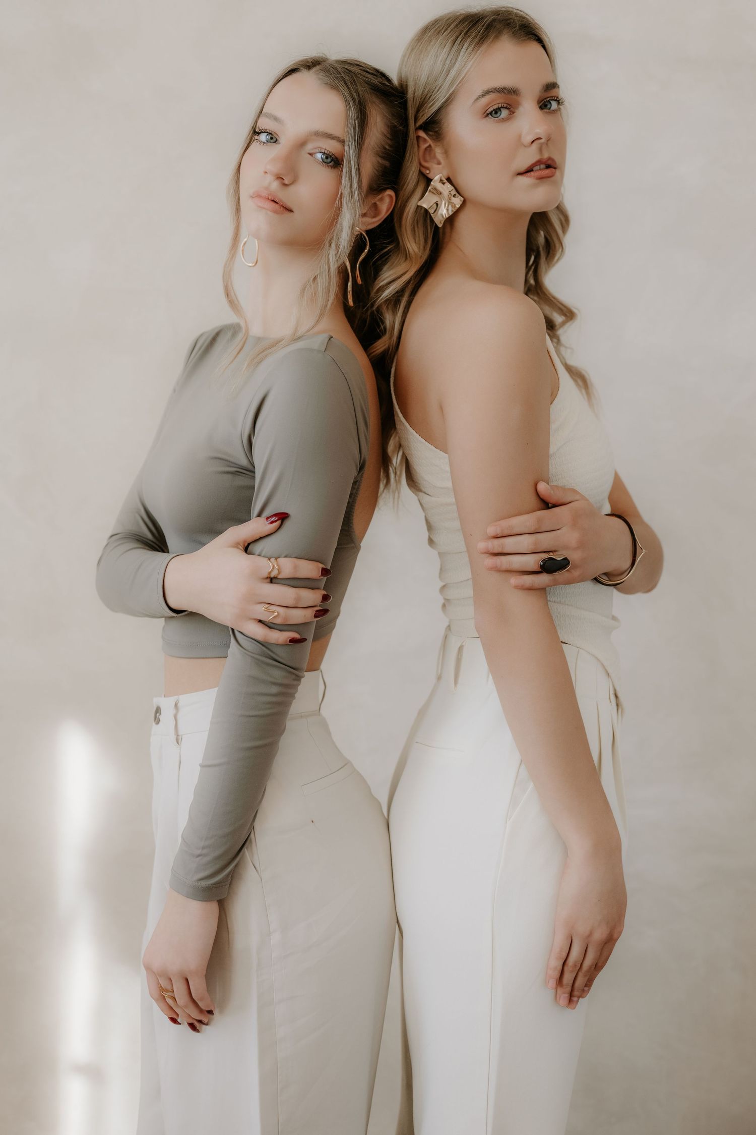 Two blonde women in neutral tones posing back-to-back against a beige studio background.