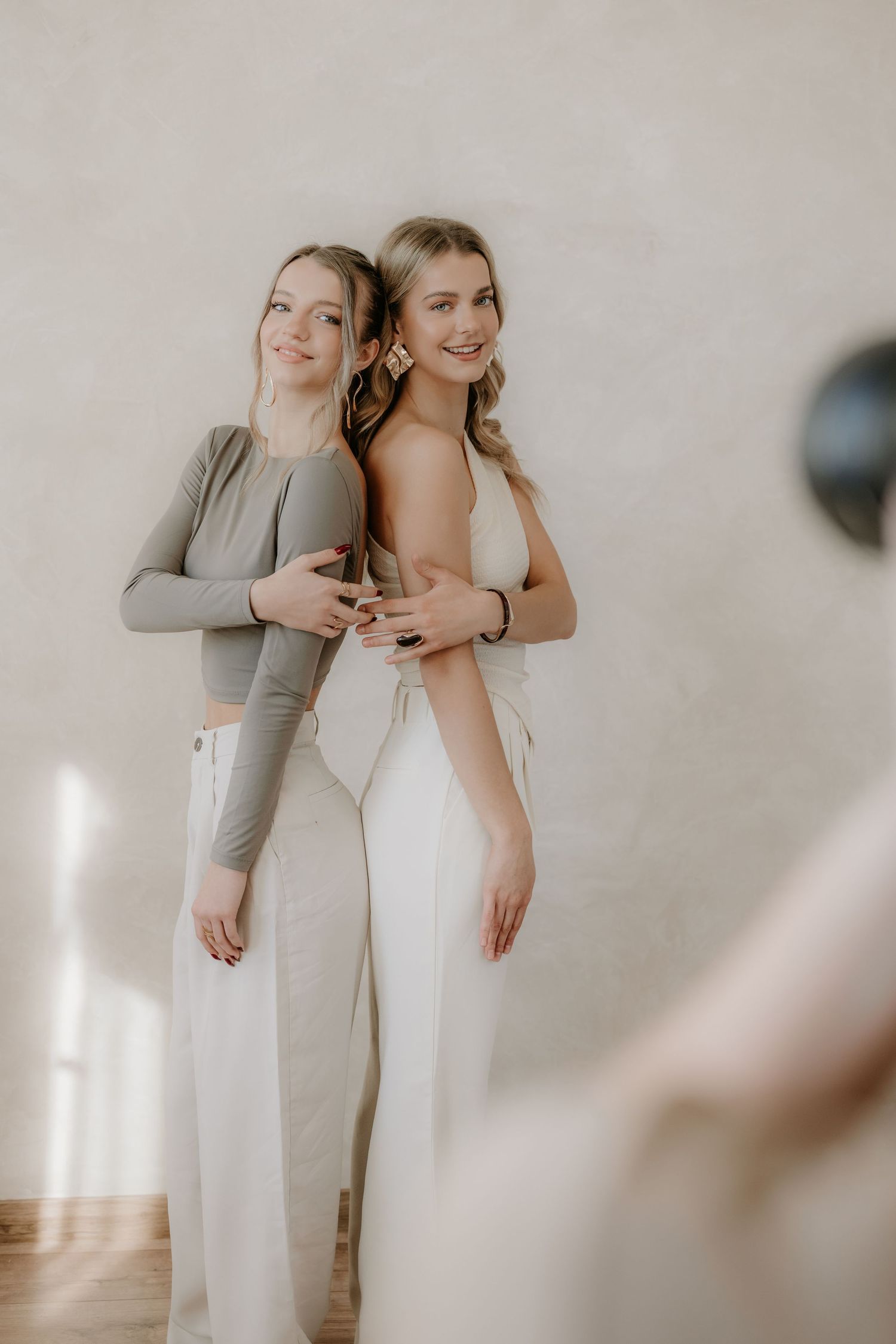 Two blonde women in neutral outfits posing back-to-back and smiling in a bright studio.