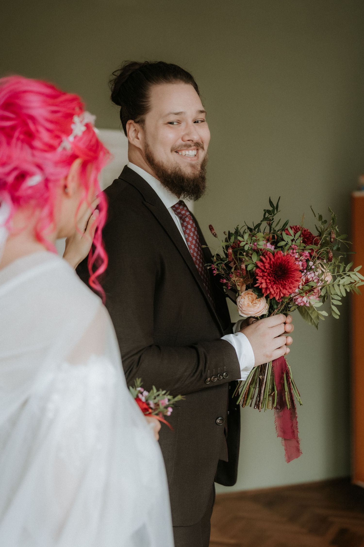 Bearded groom in dark suit smiles holding red floral bouquet facing bride with pink hair in white dress.