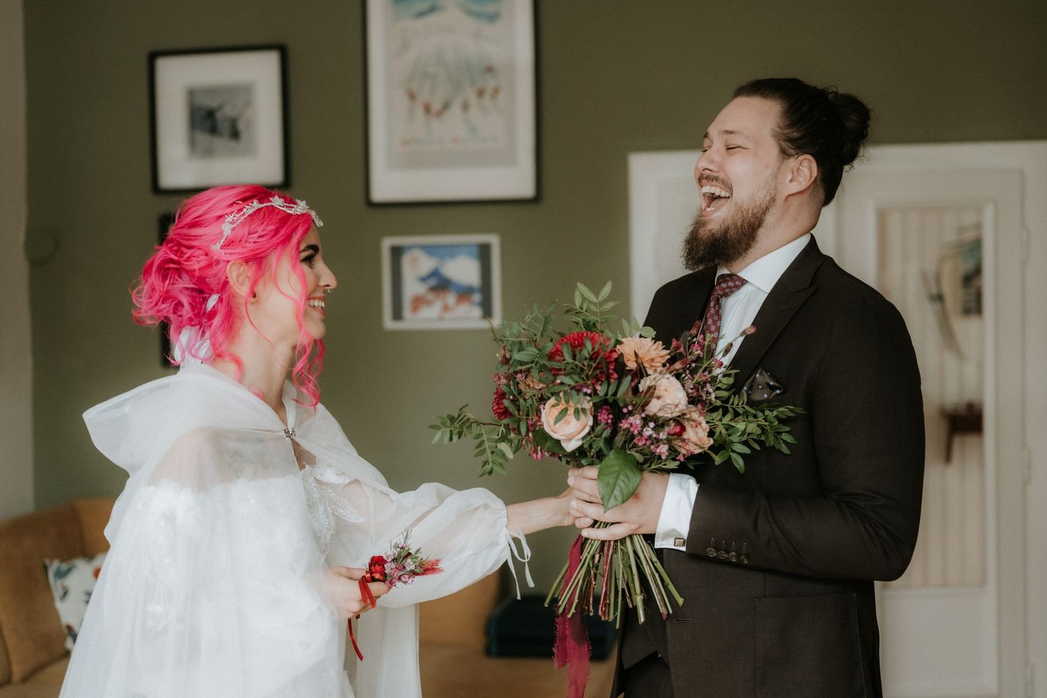 Groom in dark suit presents colorful bouquet to pink-haired bride in white dress, both smiling joyfully indoors.