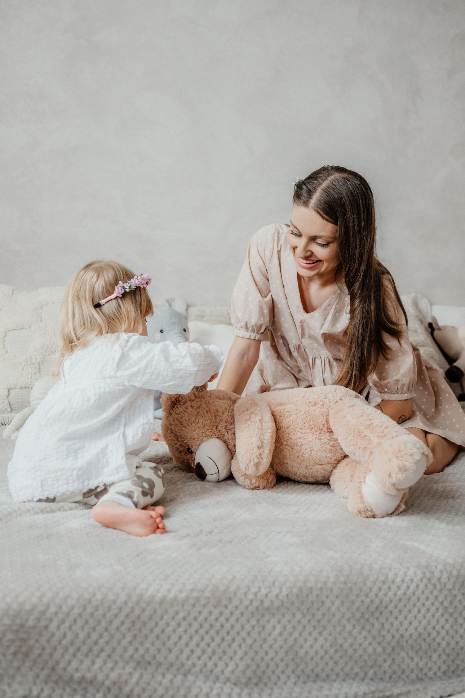Young mother smiling while playing with toddler daughter and teddy bear on a bed.