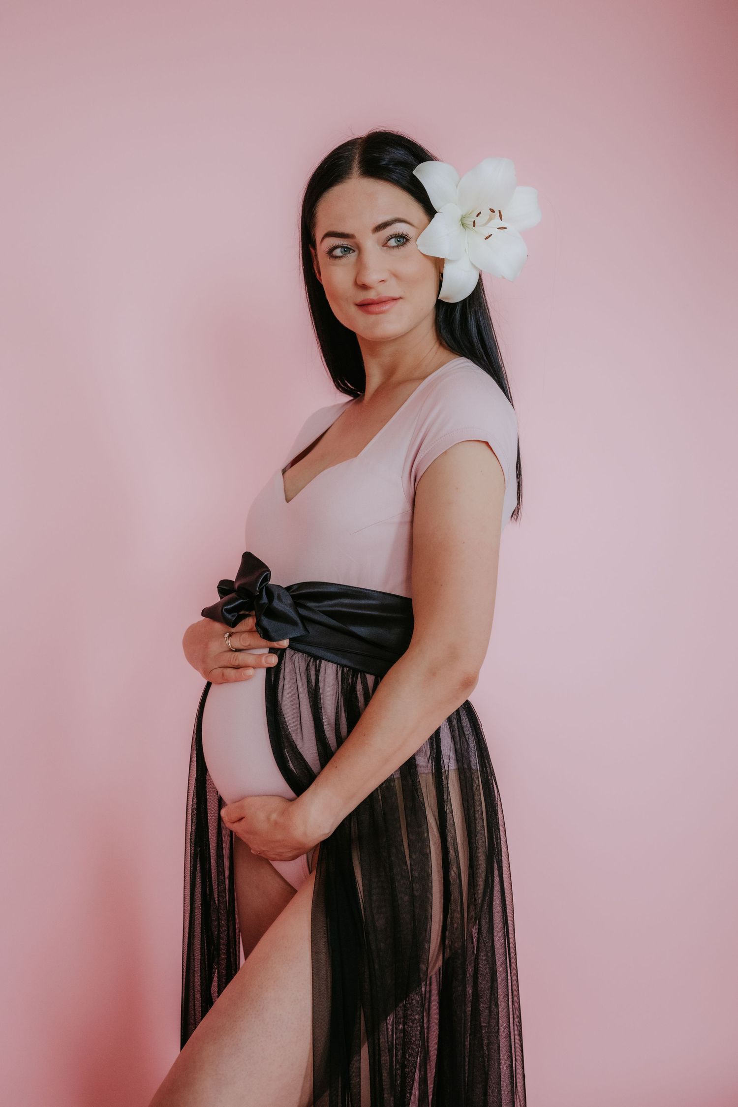 Pregnant woman in pink bodysuit with black bow and tulle skirt holding belly with flower in hair.