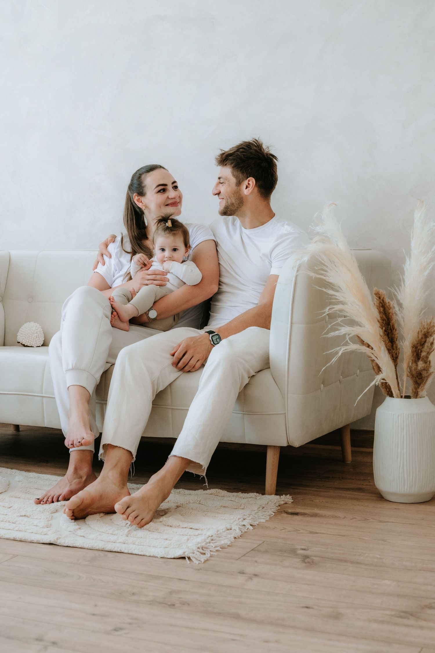 Loving young couple sitting close on a cream sofa, cradling their baby in a bright minimalist living room.