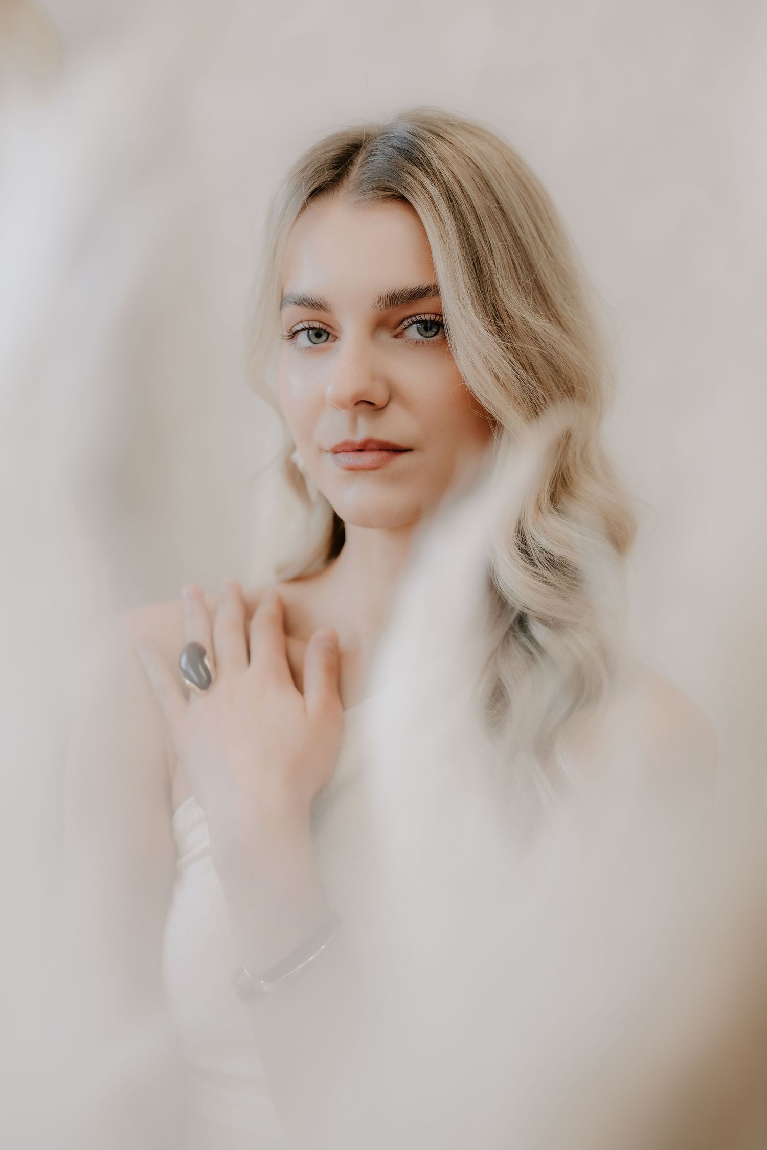 Soft-focus portrait of blonde woman with blue eyes touching her chest, wearing white top.