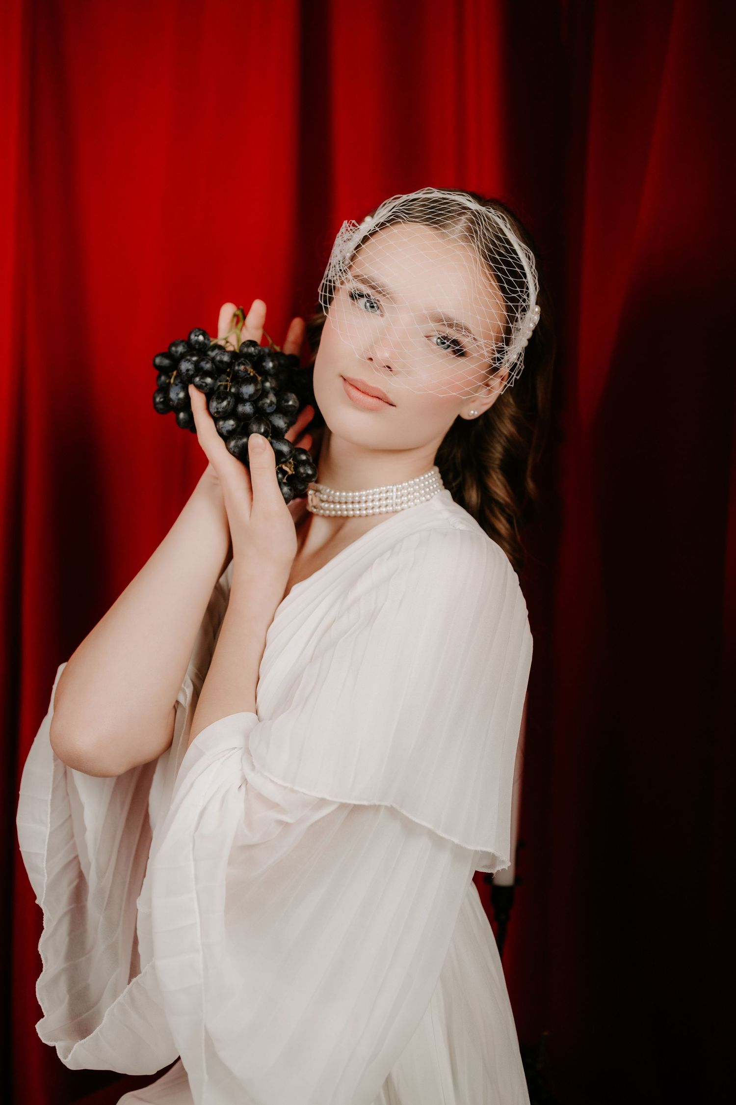 Bride in white dress holding black grapes wearing pearl choker and veil headpiece against red curtain.