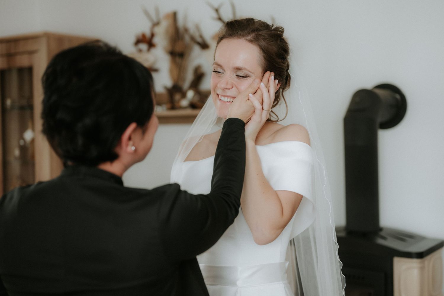 Bride in white off-shoulder gown smiles as woman adjusts her earring during wedding preparations.