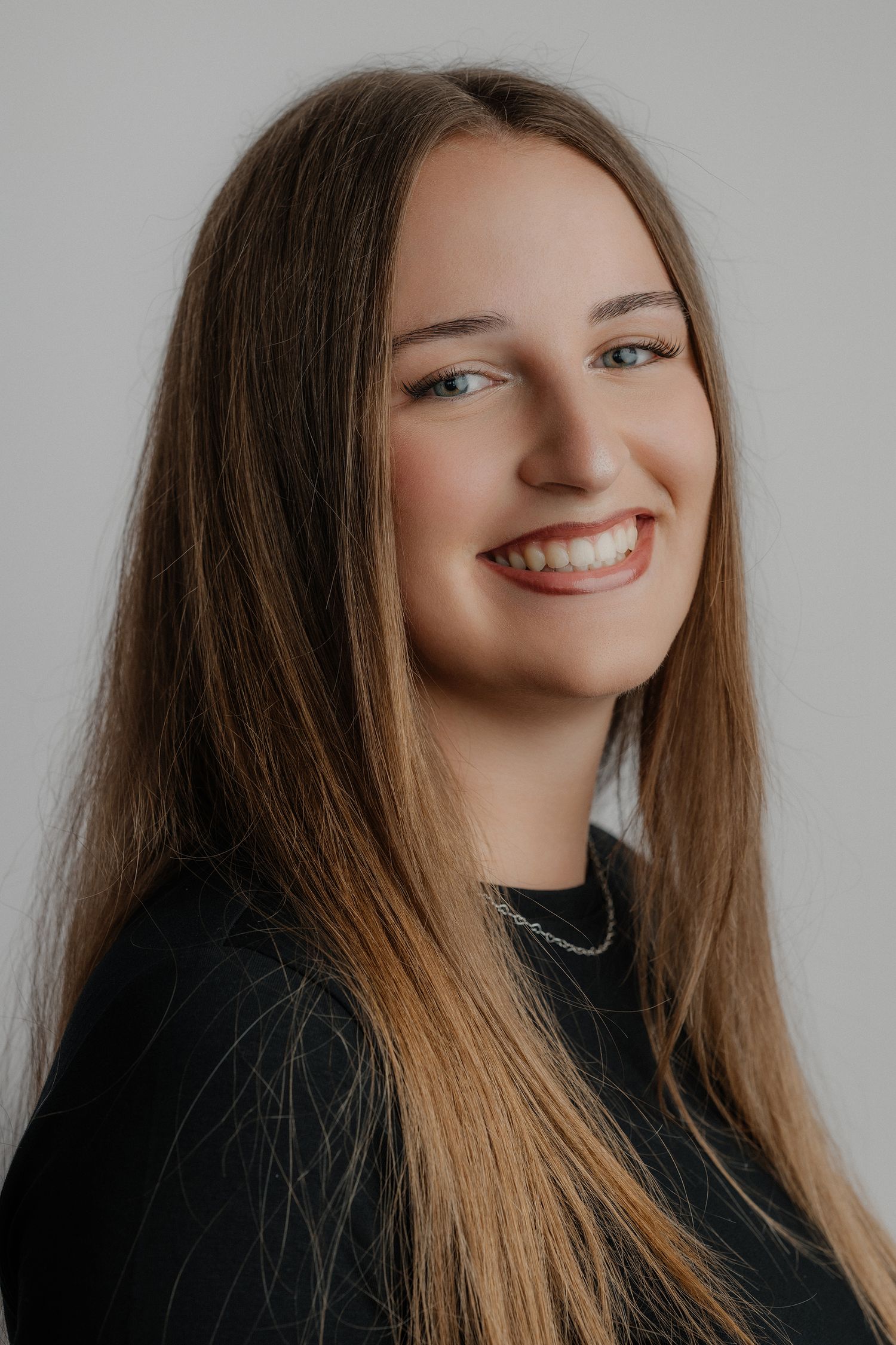 Young woman with long straight brown hair smiling broadly, wearing a black top with a chain necklace, gray background.