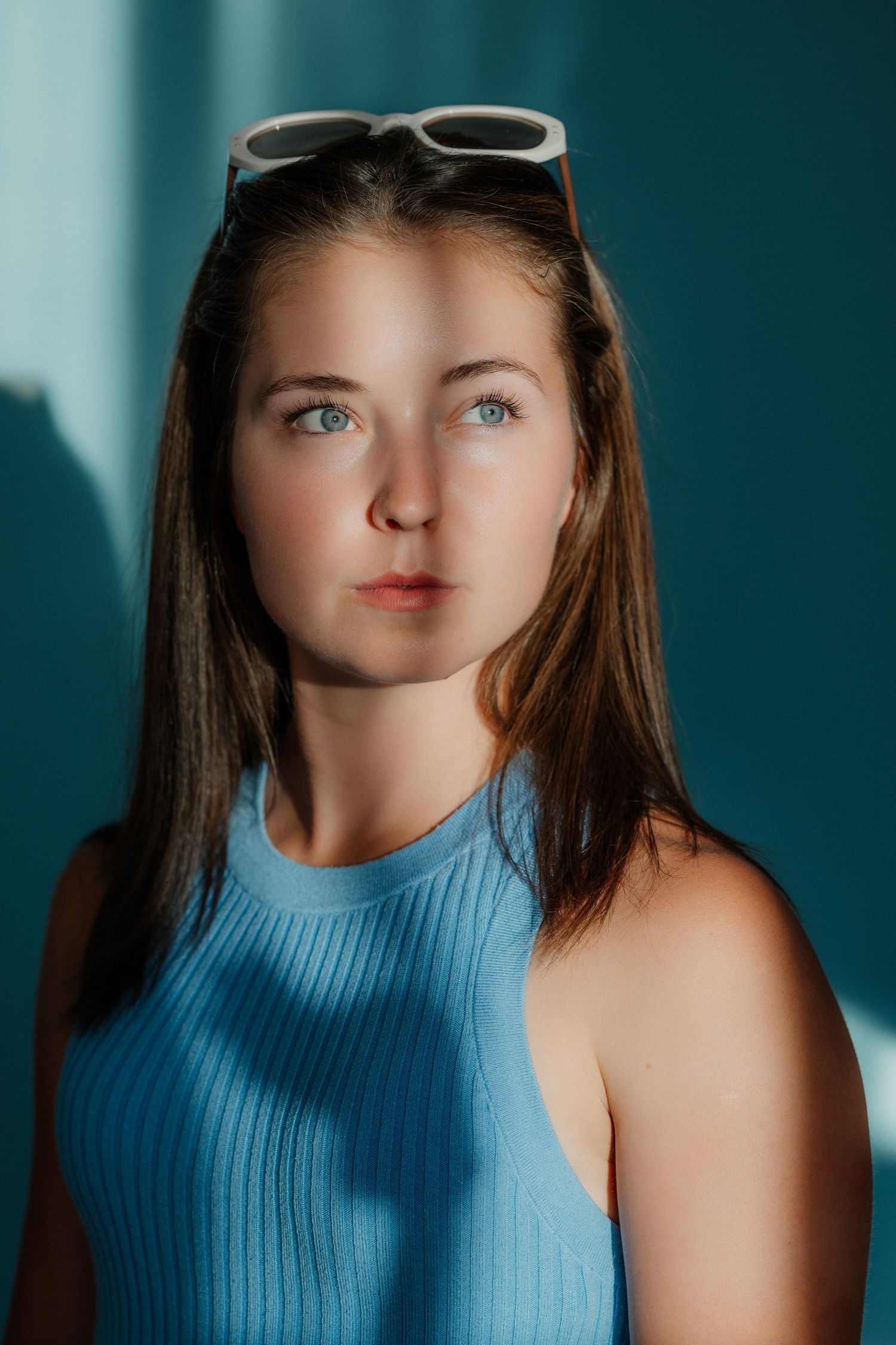 Young woman with blue eyes and sunglasses on head wearing a blue ribbed sleeveless top against a teal background.