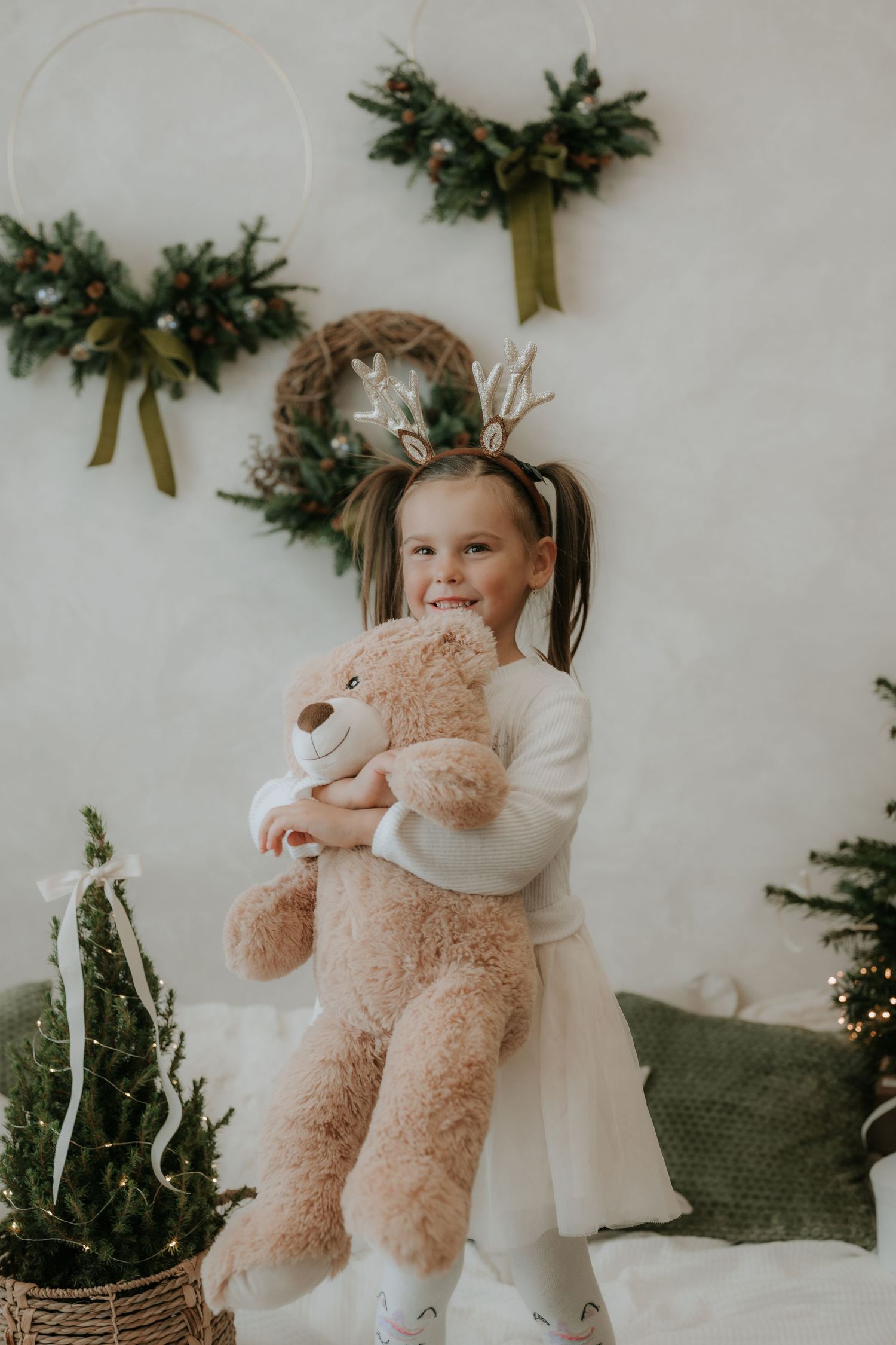 A cheerful little girl in a white dress hugs a large teddy bear wearing reindeer antlers near Christmas decorations.