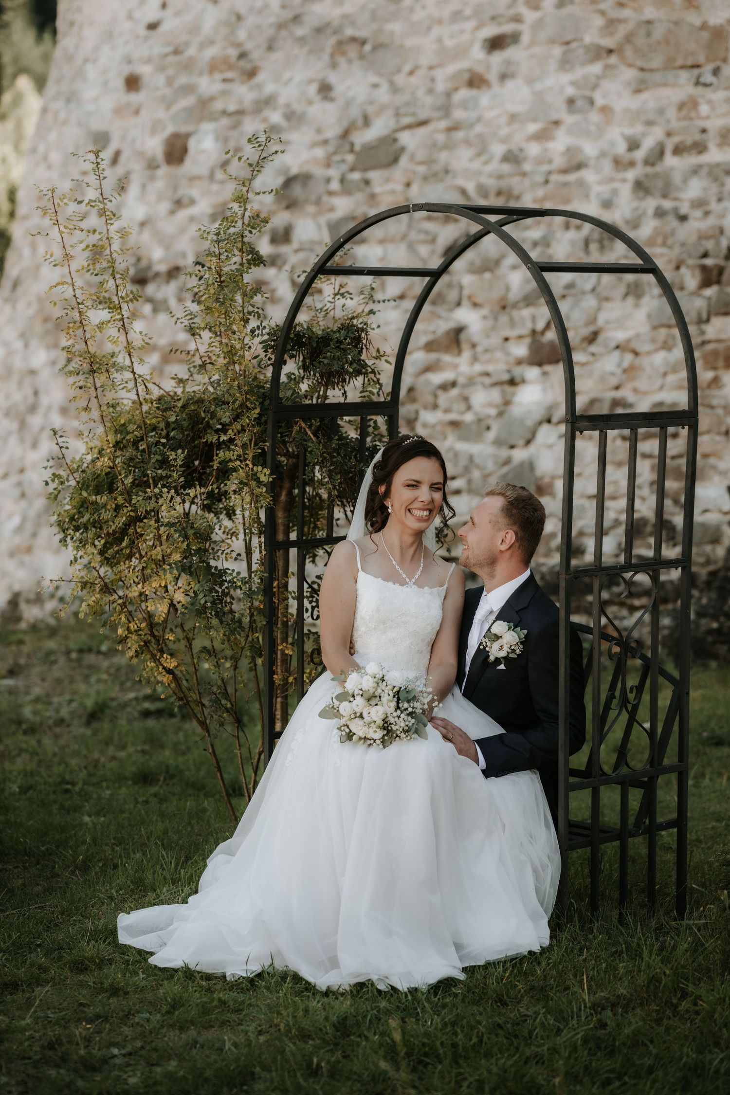Bride and groom pose under a black metal arch on green grass, with a stone wall backdrop, as she holds a white bouquet.