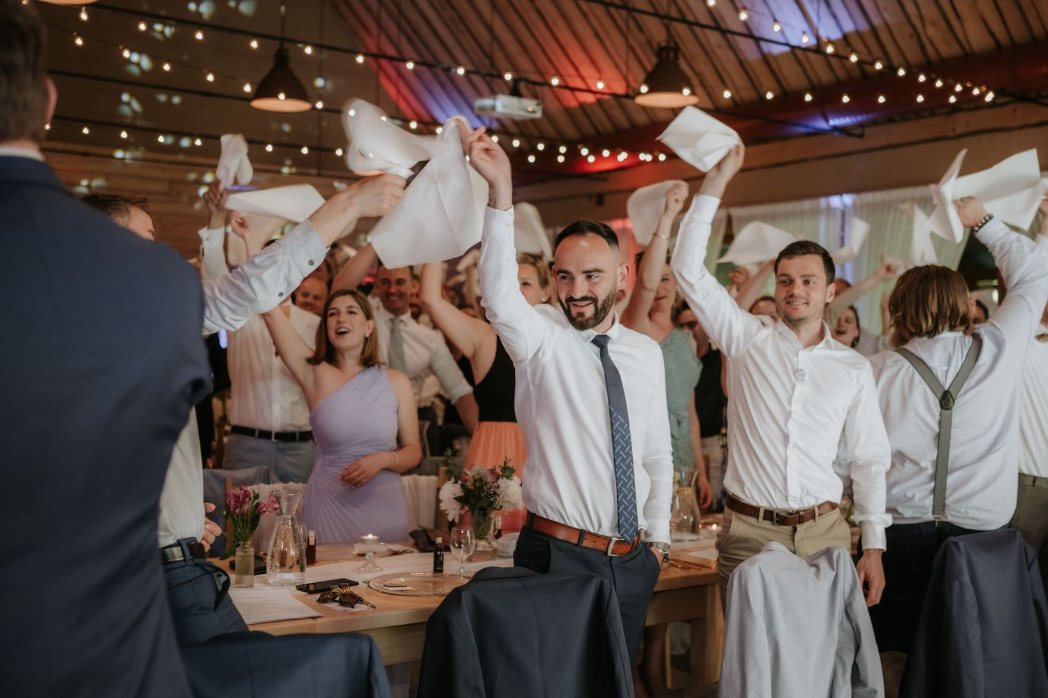 Guests wave white napkins in celebration at a lively wedding reception in a warmly lit barn venue.