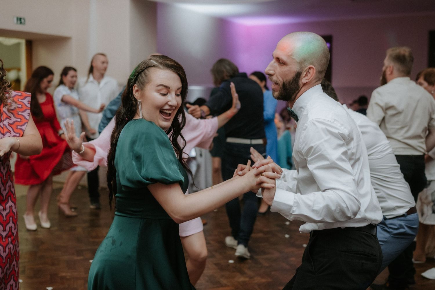 Couple dancing energetically at a wedding reception, woman in green dress, man in white shirt, colorful lights.