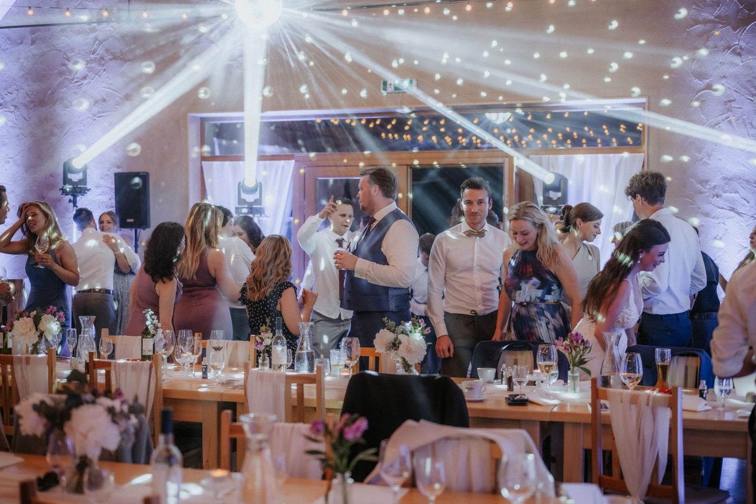 Elegant wedding reception venue with fairy lights, guests mingling around decorated tables with floral centerpieces.