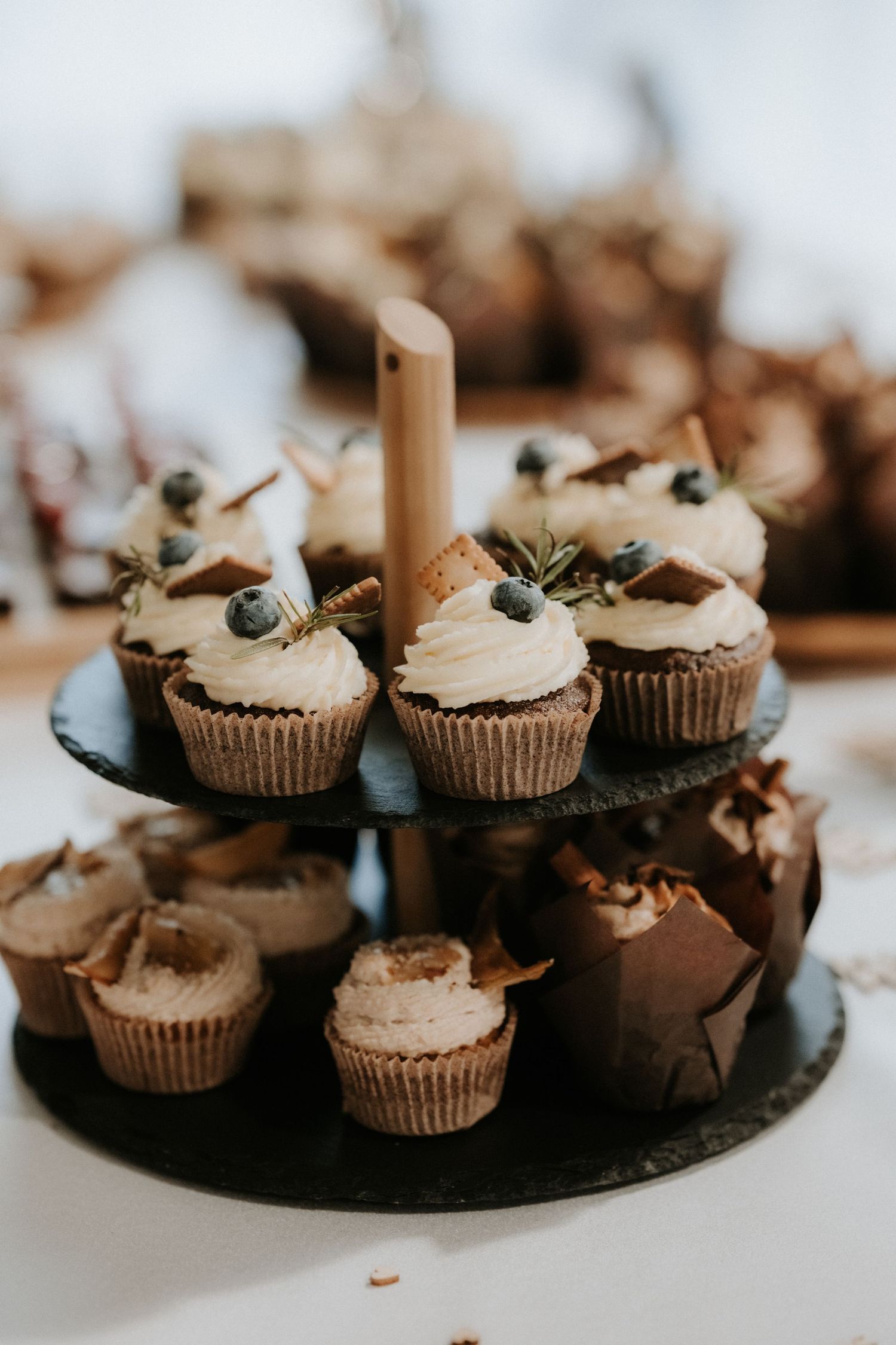 Two-tier slate stand displaying frosted cupcakes topped with blueberries and chocolate shards at a wedding dessert table.