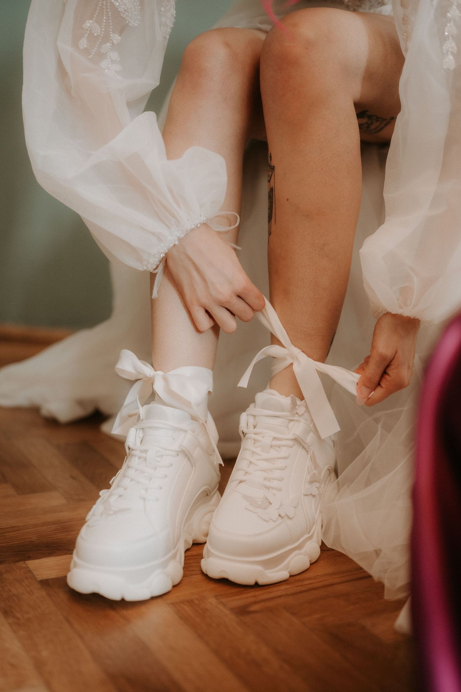 Bride in white tulle dress tying satin ribbon bows on chunky white sneakers on wooden floor.