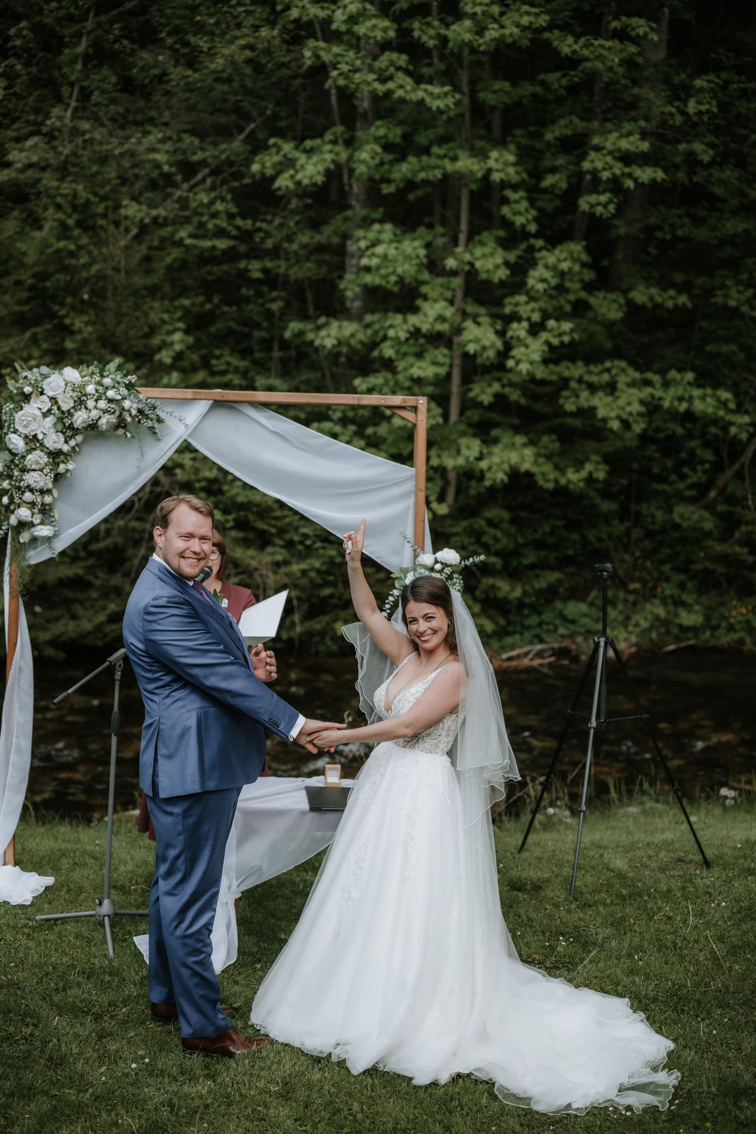 Joyful bride and groom celebrate at outdoor wedding ceremony with floral arch, white draping, and lush green forest backdrop.