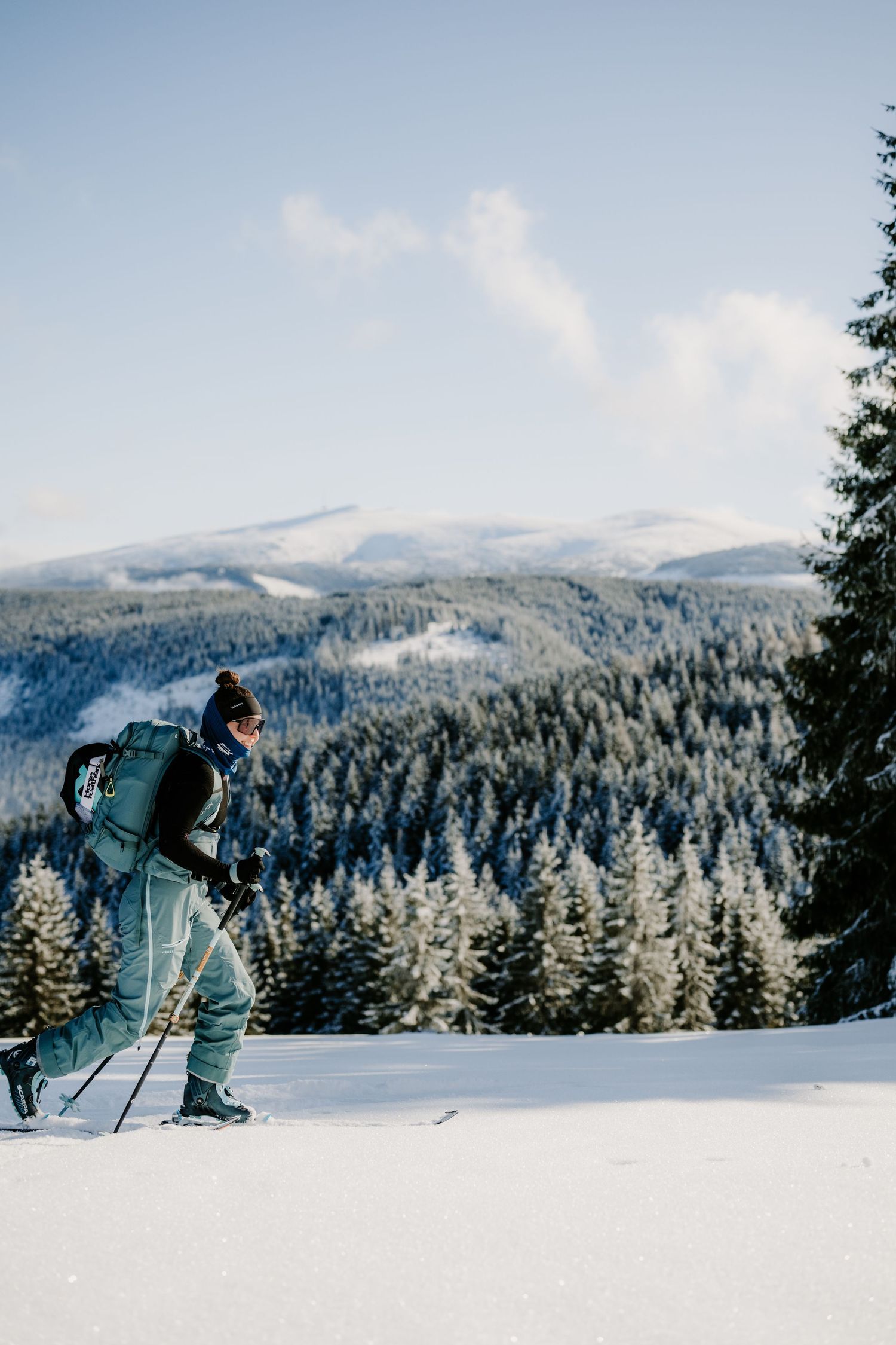 A backcountry skier with a large pack traverses a snowy slope with forested mountains in the background.