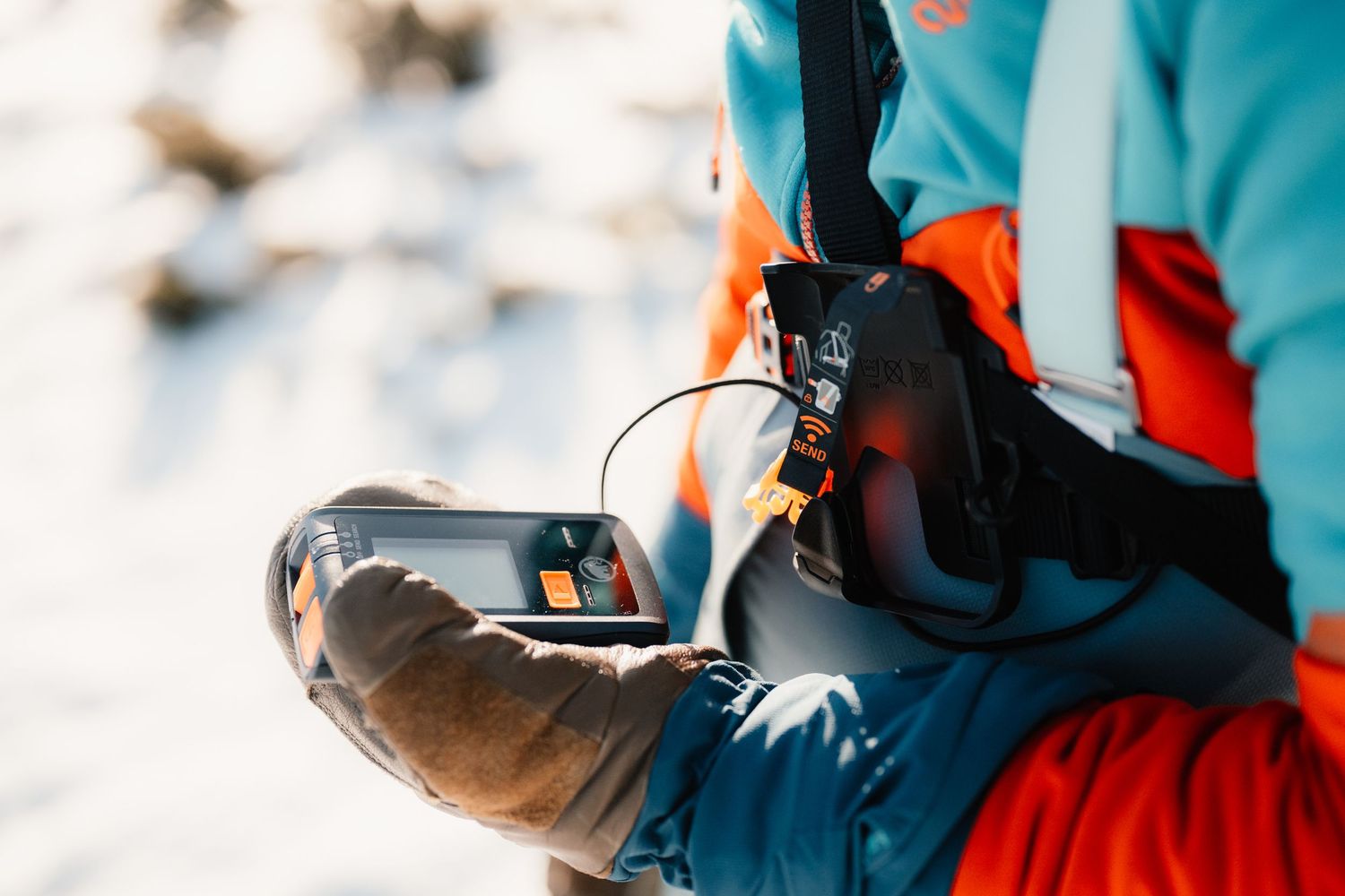 Skier in teal jacket using avalanche safety device with orange beacon in snowy mountain environment.