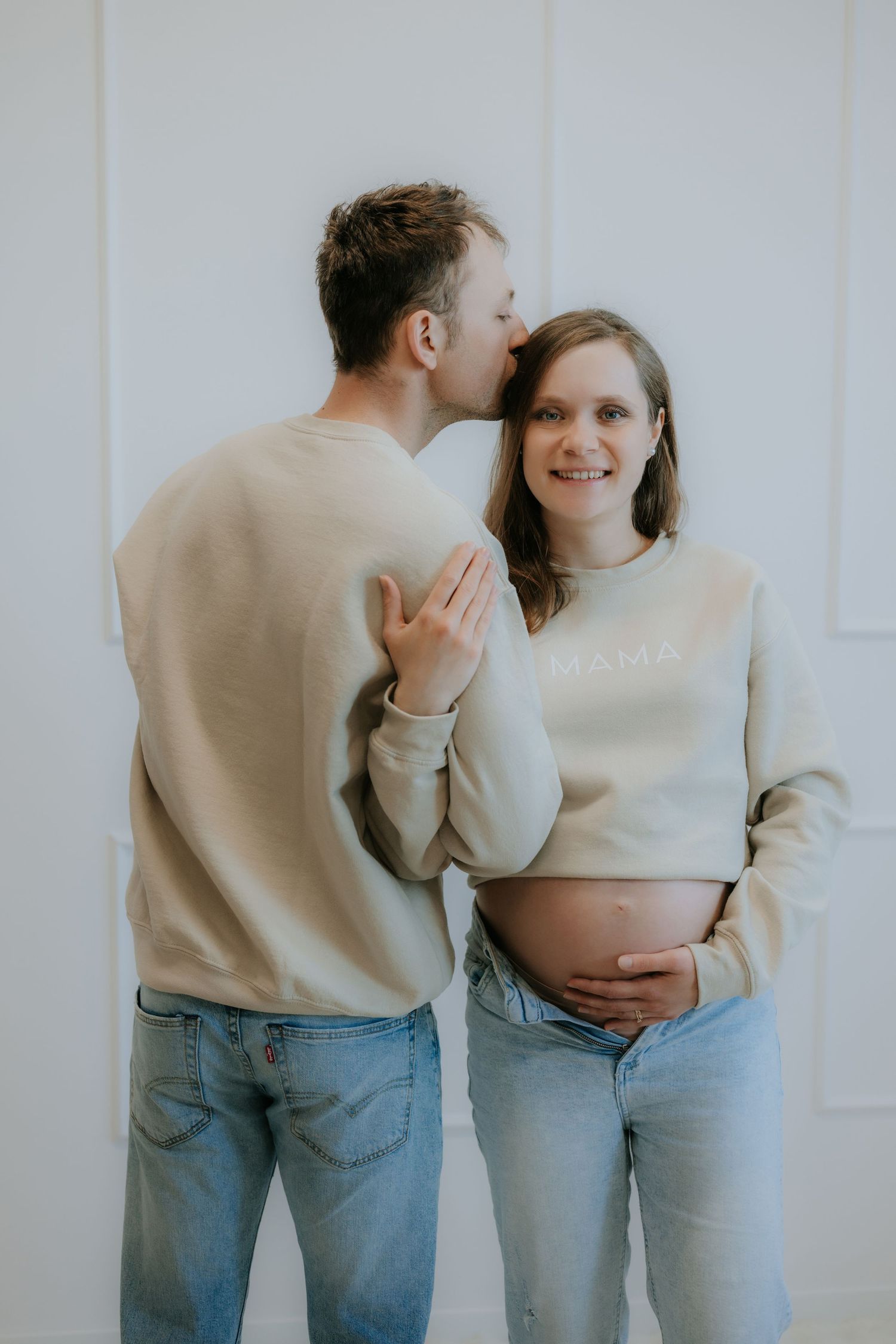 Expectant couple in matching beige sweatshirts, man kissing pregnant womans cheek tenderly.