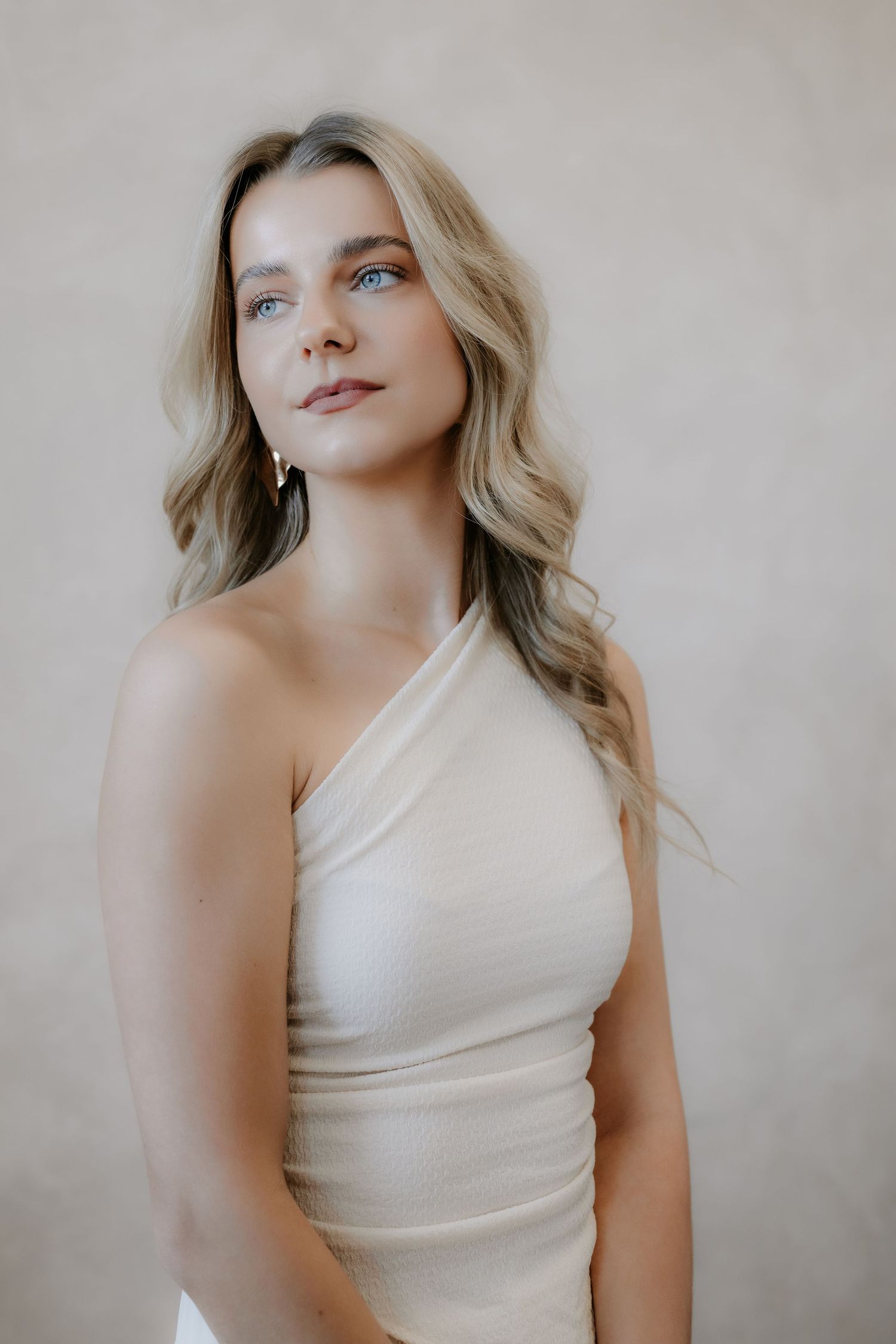 Blonde woman in elegant cream one-shoulder top posing gracefully against neutral background.