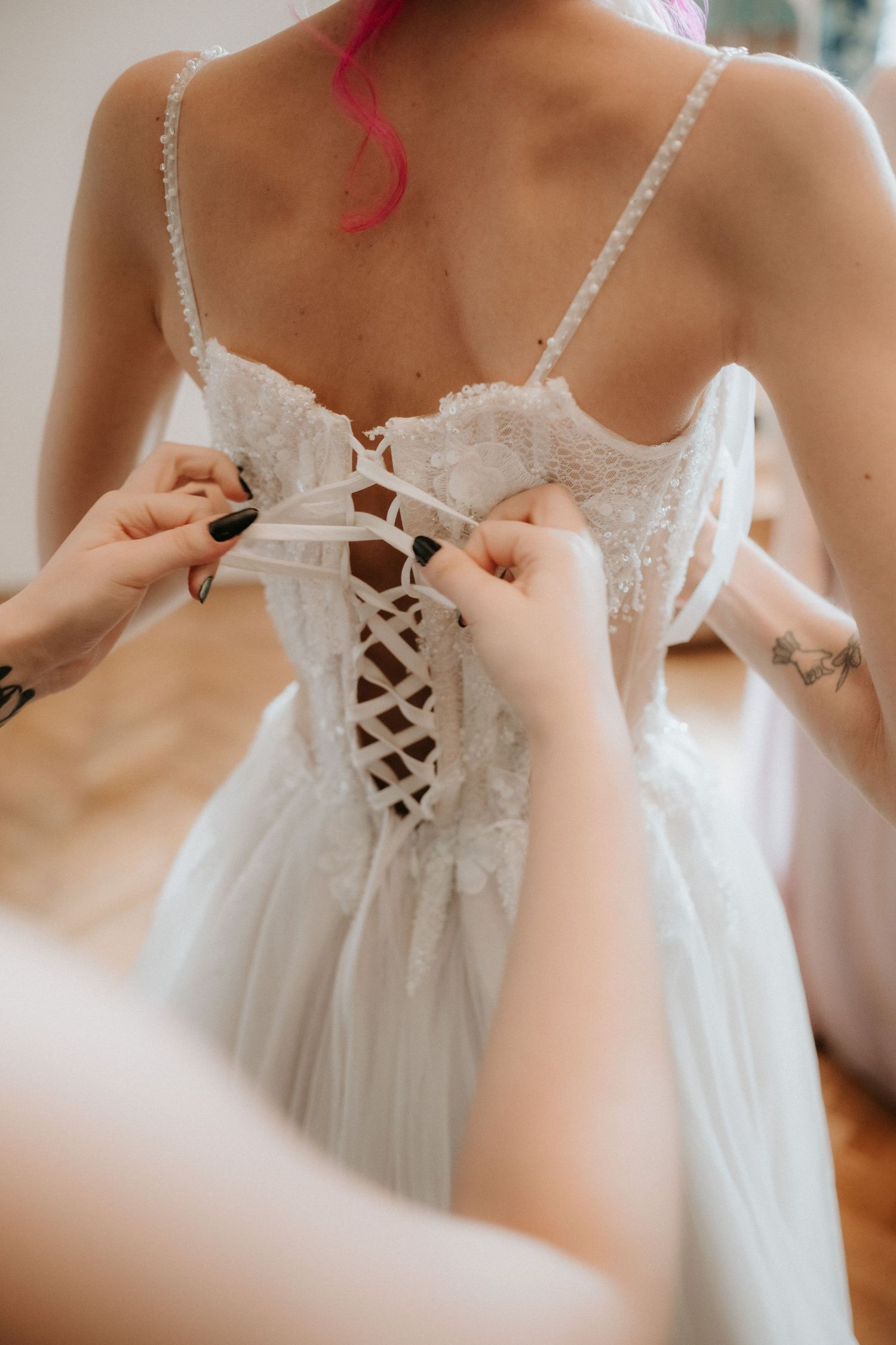 Bridesmaid lacing up corset back of white lace wedding dress on pink-haired bride.