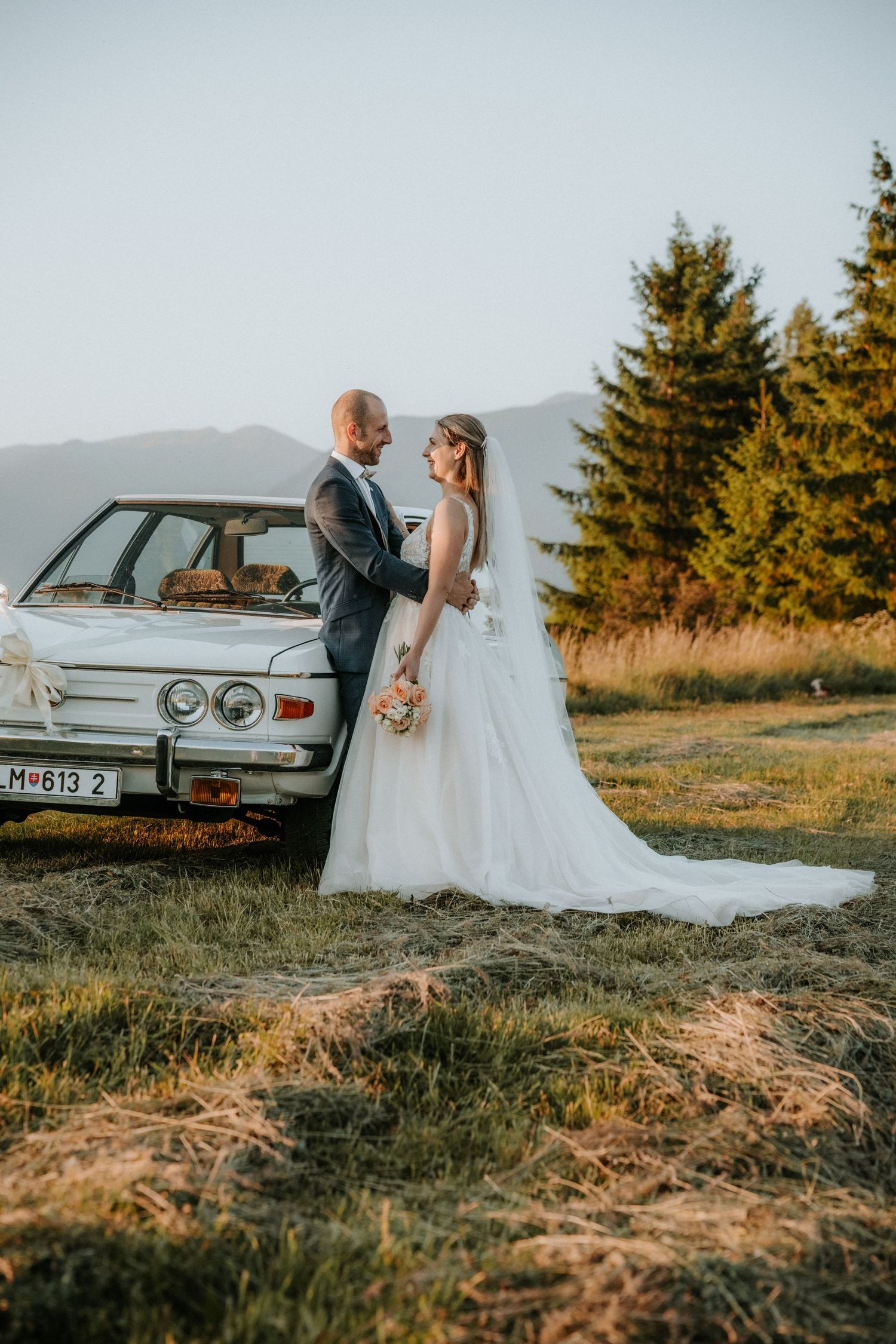 Bride and groom stand together by a vintage white car in a scenic mountain meadow at golden hour.