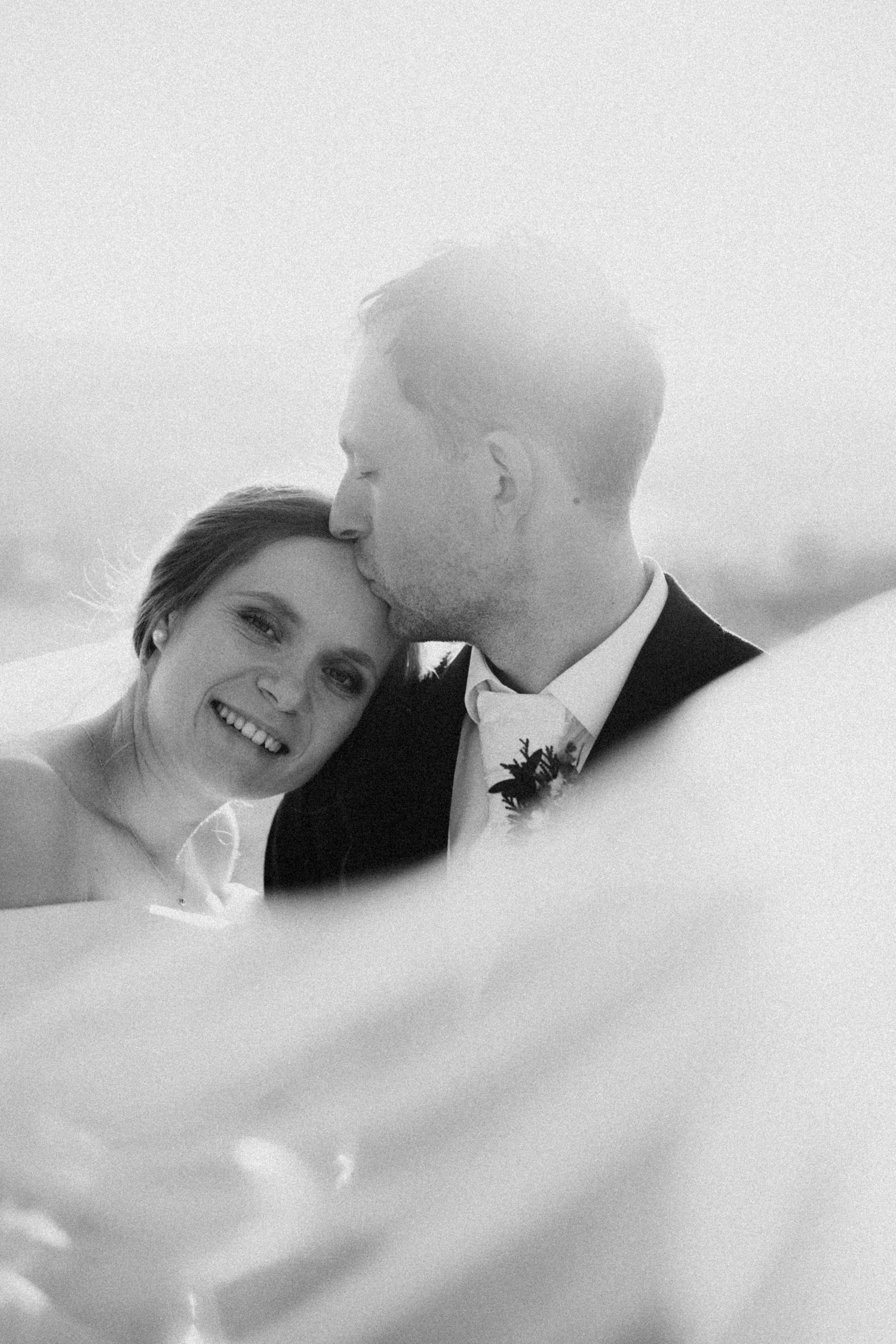 Black and white wedding photo of smiling bride and groom sharing a tender kiss, framed by flowing veil.