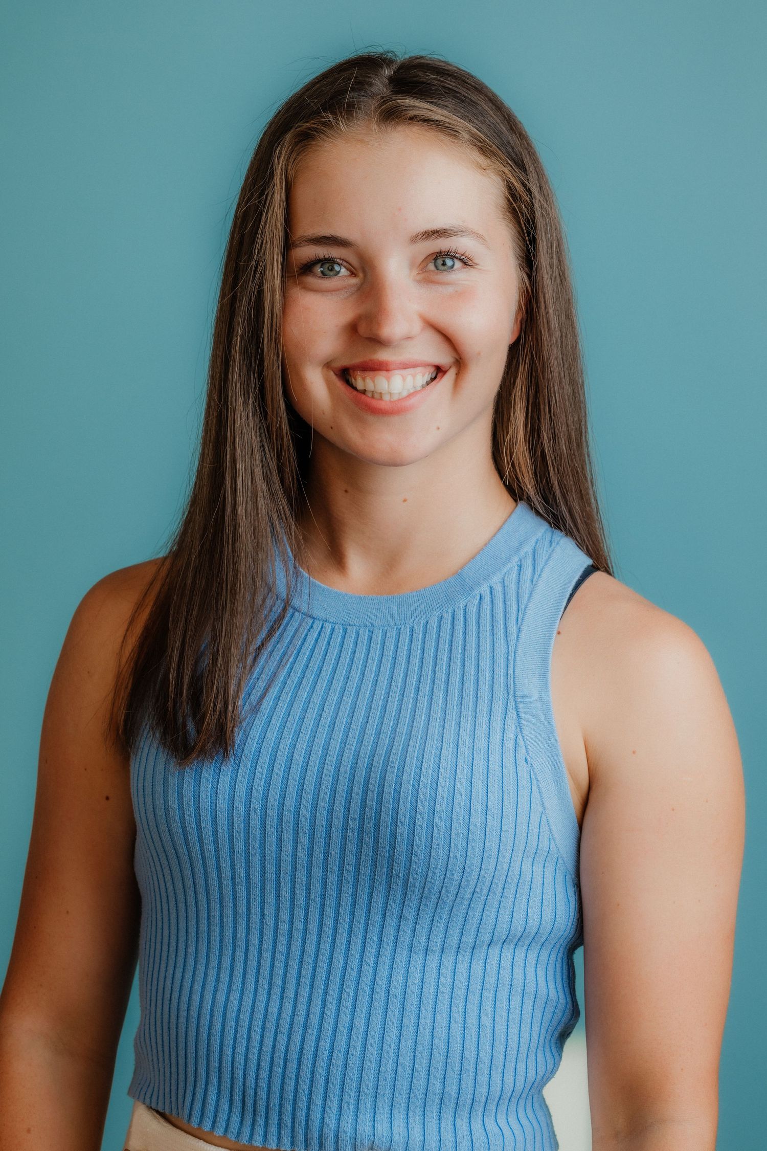 Smiling young woman with brown hair wearing a blue ribbed crop top against a teal background.