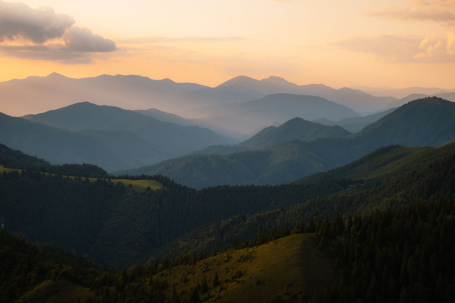 Layered mountain ridges covered in forests at sunset with dramatic orange and blue sky tones.