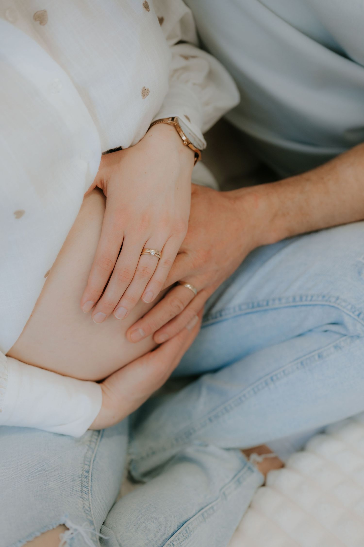 Close-up of pregnant woman and partner holding hands on baby bump with gold ring and watch.