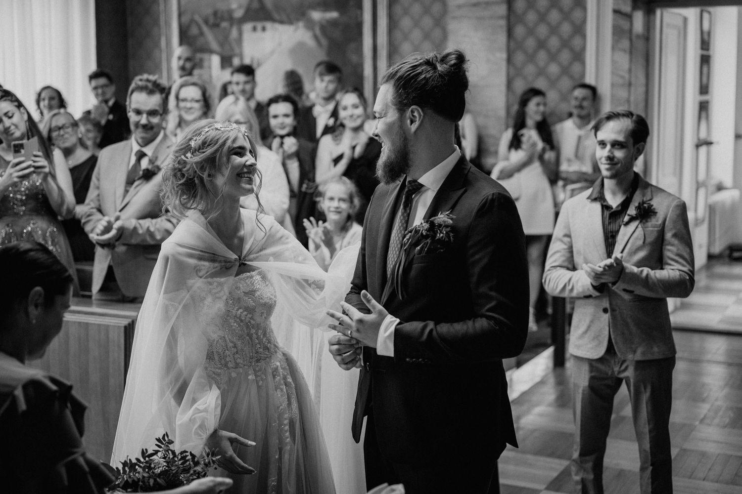 Black and white wedding ceremony photo of bride and groom exchanging vows surrounded by guests.