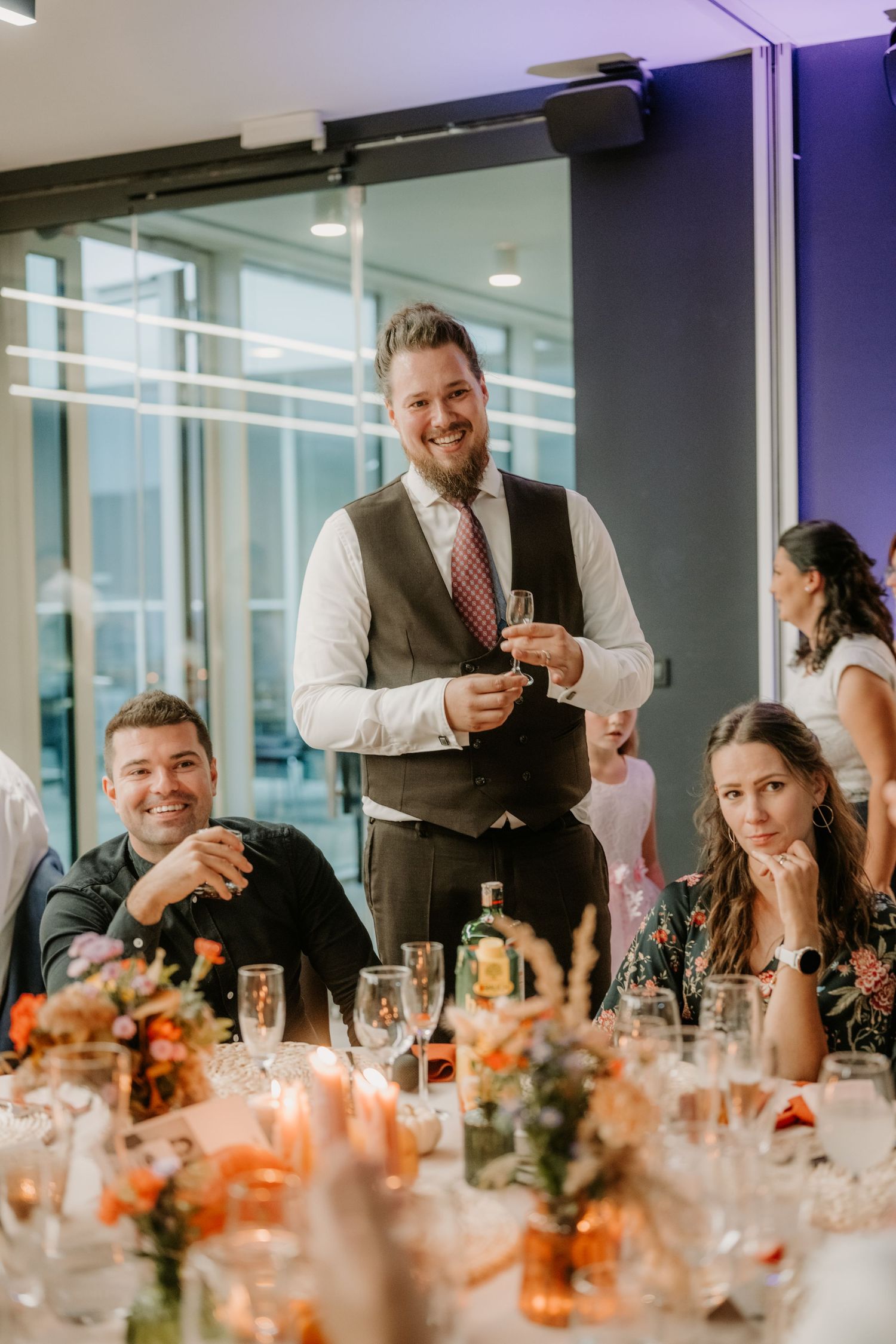 Man in vest giving a wedding speech at a elegantly decorated reception table with guests smiling.