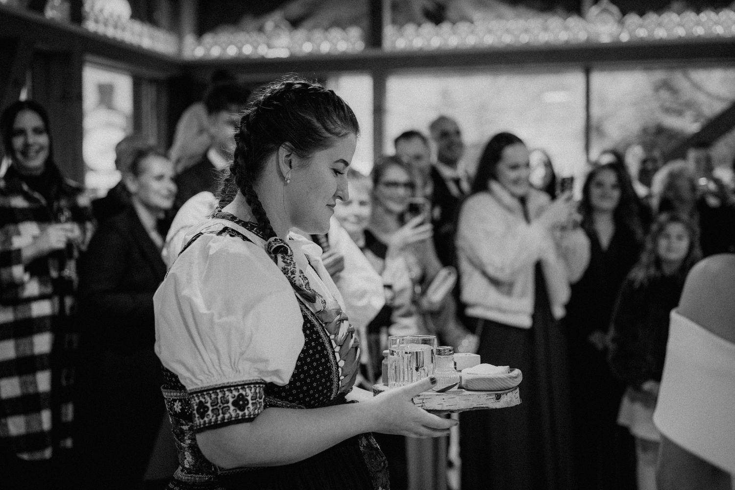 Woman in traditional folk costume serving drinks on a tray at a festive gathering, black and white photo.