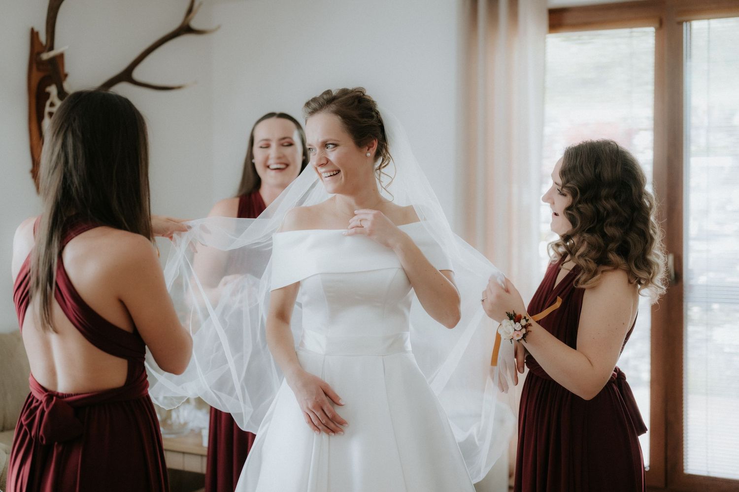 Bride smiling in white wedding gown as bridesmaids in burgundy dresses adjust her veil indoors.