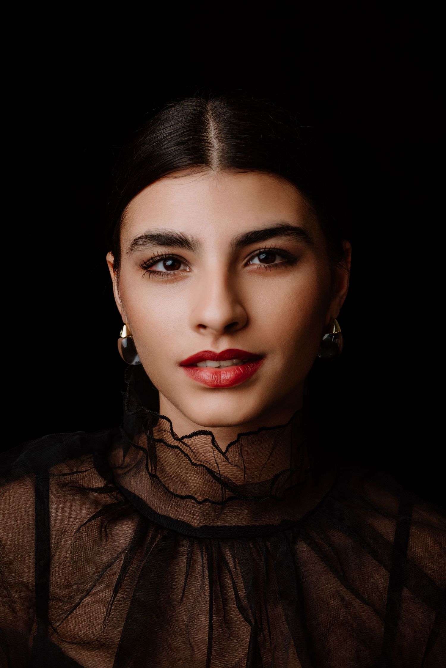 Elegant woman with red lips and gold earrings wearing a sheer black high-neck top against dark background.