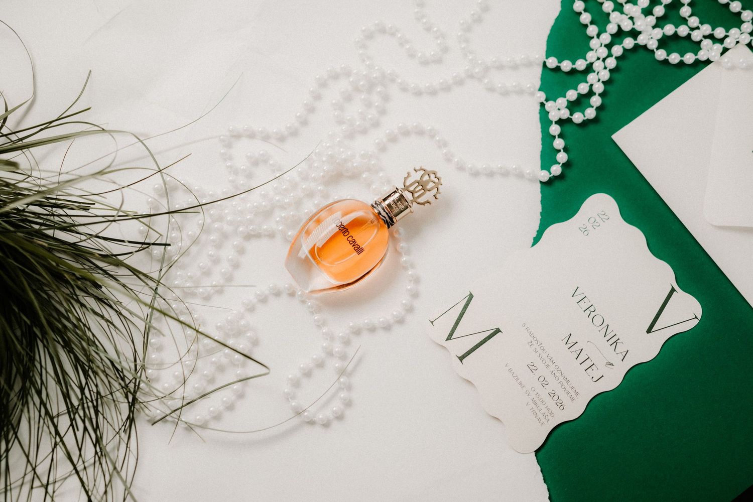 Close-up flat lay of an amber perfume bottle with pearl necklace and wedding invitation on green surface.